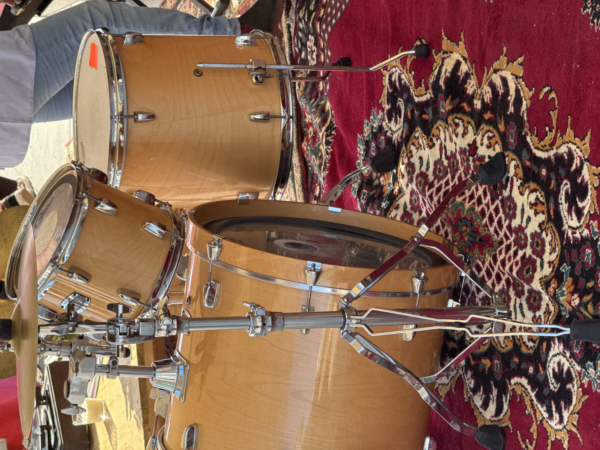 The image displays a drum set with a natural wood finish, resting on a red oriental rug. The drums are angled, suggesting they are part of a musical performance or setup. Several chrome stands and clamps are visible, holding the drums and potentially other cymbal stands. In the background, a person's legs, clad in blue jeans, are visible, indicating a person is present near the drums. The lighting suggests it could be daytime, with natural light illuminating the scene. The overall impression is that of a casual musical gathering or practice session. The setting appears to be outdoors, possibly in a courtyard or a public space, given the natural light and the presence of a rug on a surface that could be pavement or a wooden deck. There is no visible text in the image.