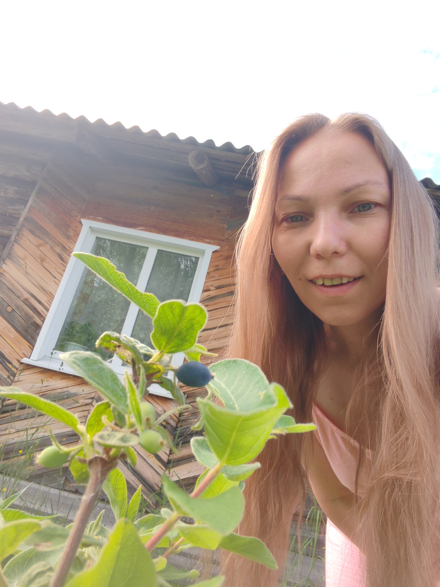 A woman with long, light brown hair is taking a selfie outdoors in front of a wooden building. She is smiling and looking at the camera. In the foreground, a plant with green leaves and dark berries is visible. The wooden building appears to be a house or shed with a corrugated roof and a window. The scene is likely in a rural or suburban setting. The overall lighting suggests it is daytime, possibly late afternoon due to the warm tones. The specific location in the city of Чухлэм, Russia, is not visually identifiable from the image.