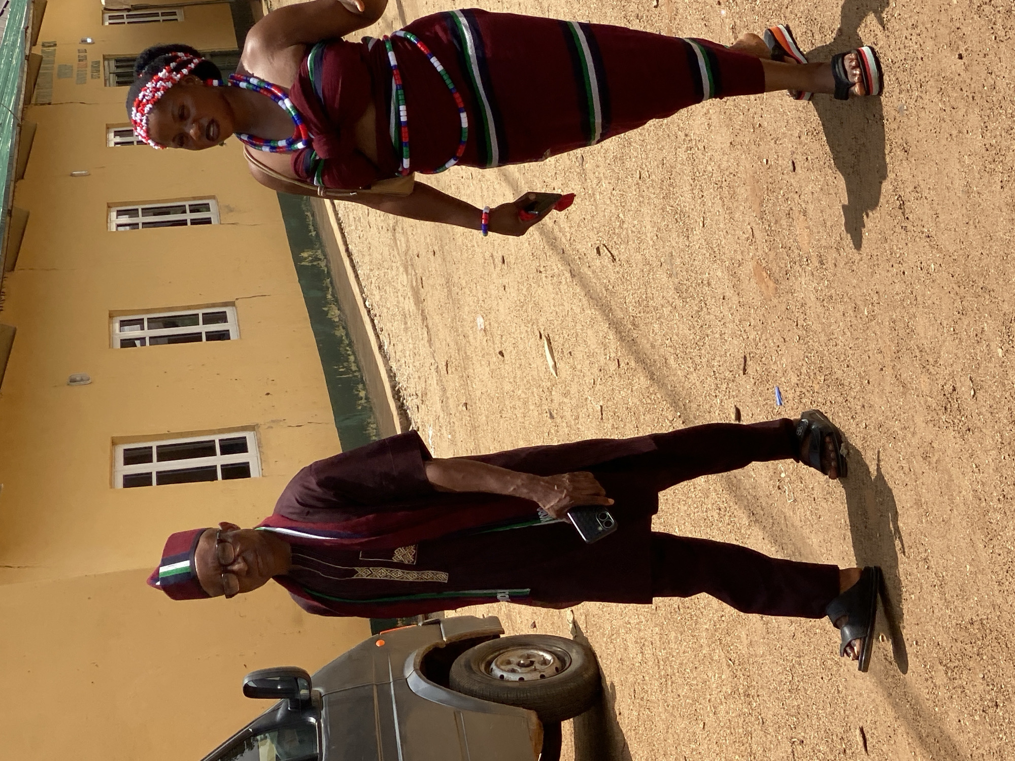 A man and woman stand outside in front of a light yellow building. The man wears a dark brown suit with a matching hat. The woman wears a burgundy dress with stripes and beaded jewelry. Both hold cell phones. The location is Maigizo, Nigeria. A gray car is partially visible near the man.