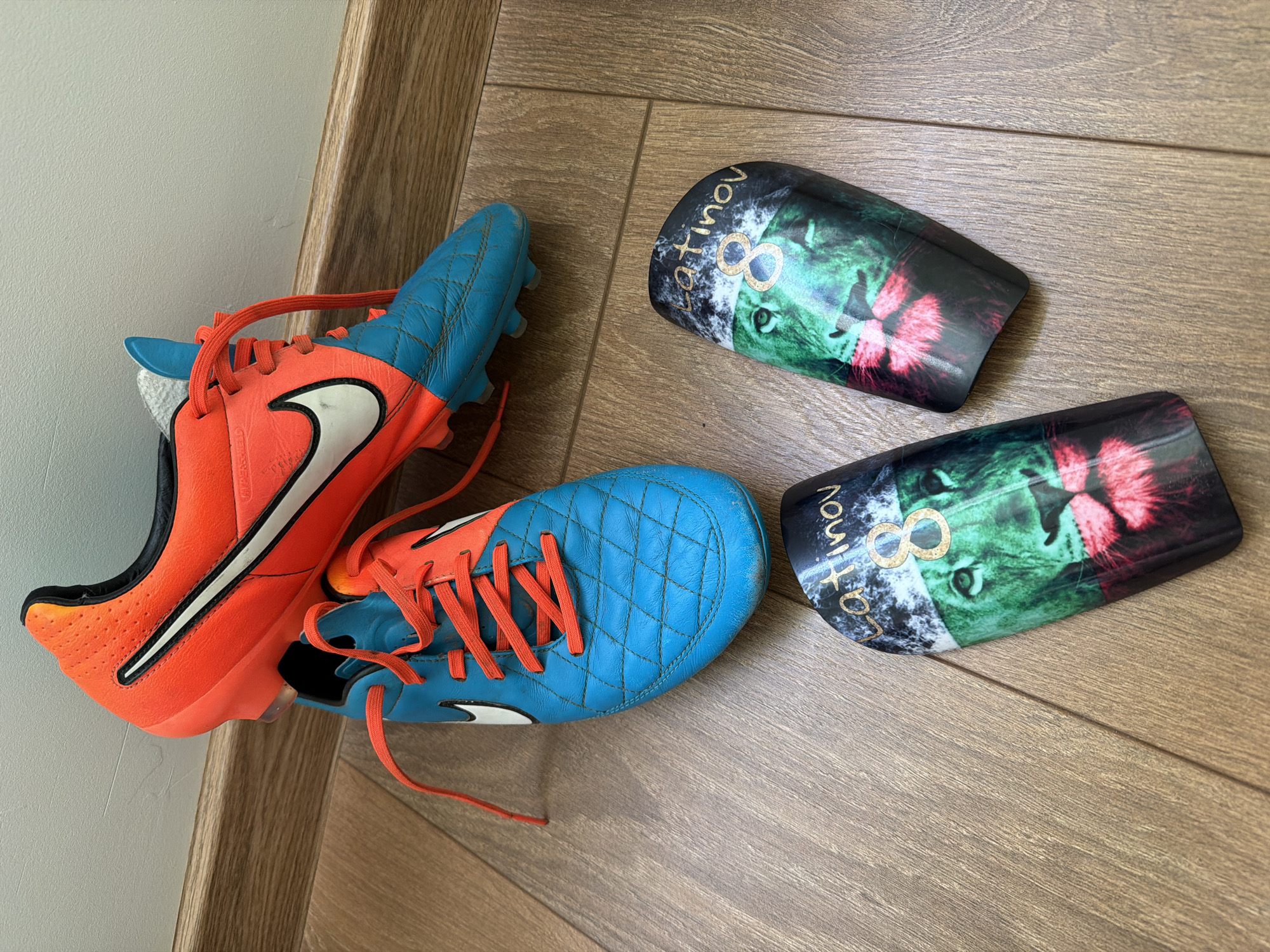 The scene depicts a pair of Nike soccer cleats in orange and blue, accompanied by two shin guards with lion imagery, lying on a wood-patterned floor. The cleats are laced with orange laces and show signs of use. The shin guards feature a lion face split into green and red, with the name "Latinov" and the number 8 printed on them. The setting appears to be indoors, possibly a residence, but the specific location is unknown.