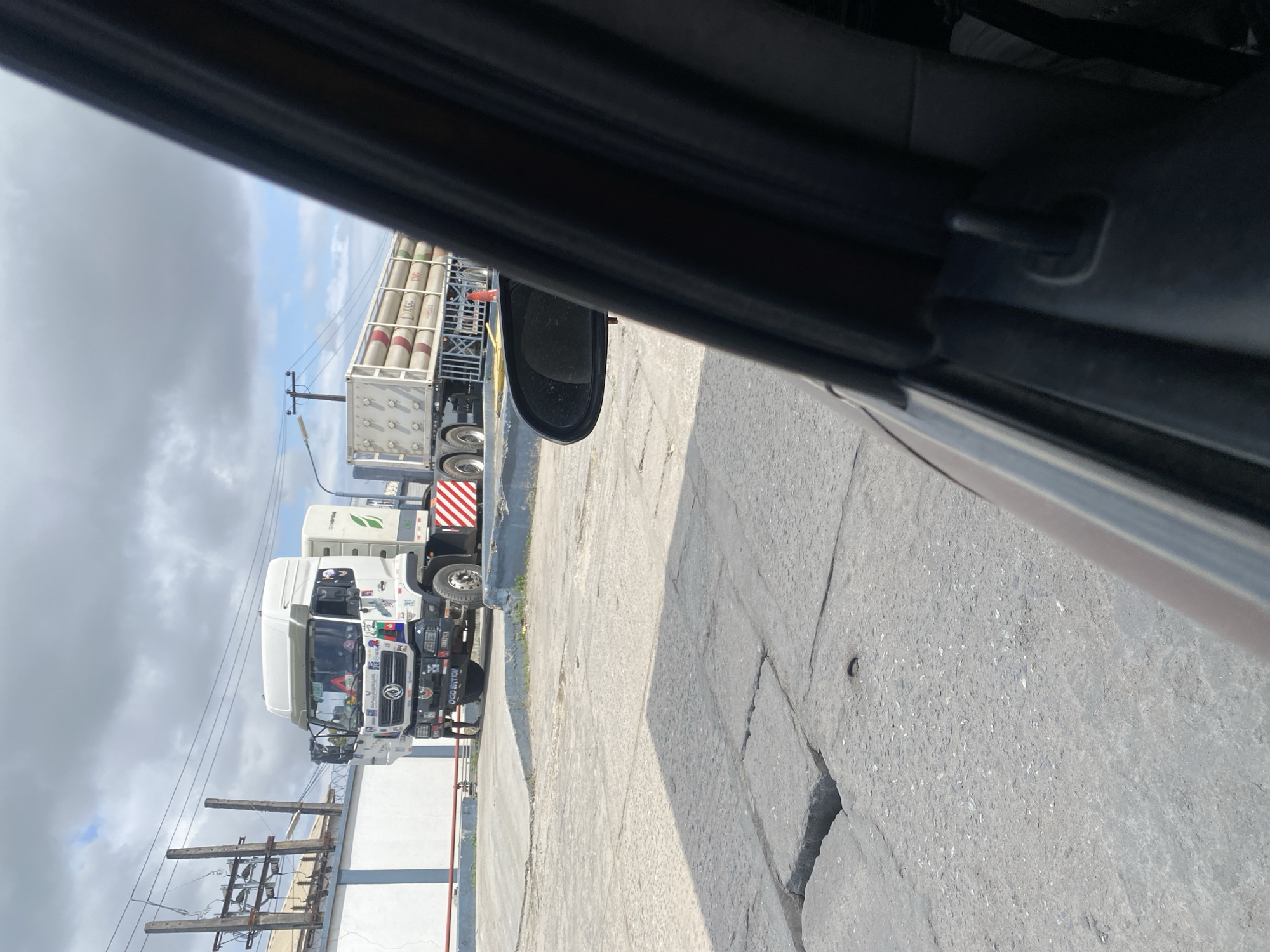 A white truck with a flatbed trailer loaded with cylindrical tanks is parked on a paved surface in Ajah, Nigeria. The truck has a white cab with various stickers and emblems on its exterior. A person stands near the front of the truck. The trailer is secured with a metal railing. In the background, there is a white building with electrical equipment on poles and overhead wires. The scene is outdoors under a cloudy sky. A car's interior, including a side mirror, is visible in the foreground, suggesting the photo was taken from a vehicle. The ground is made of large, cracked concrete slabs.