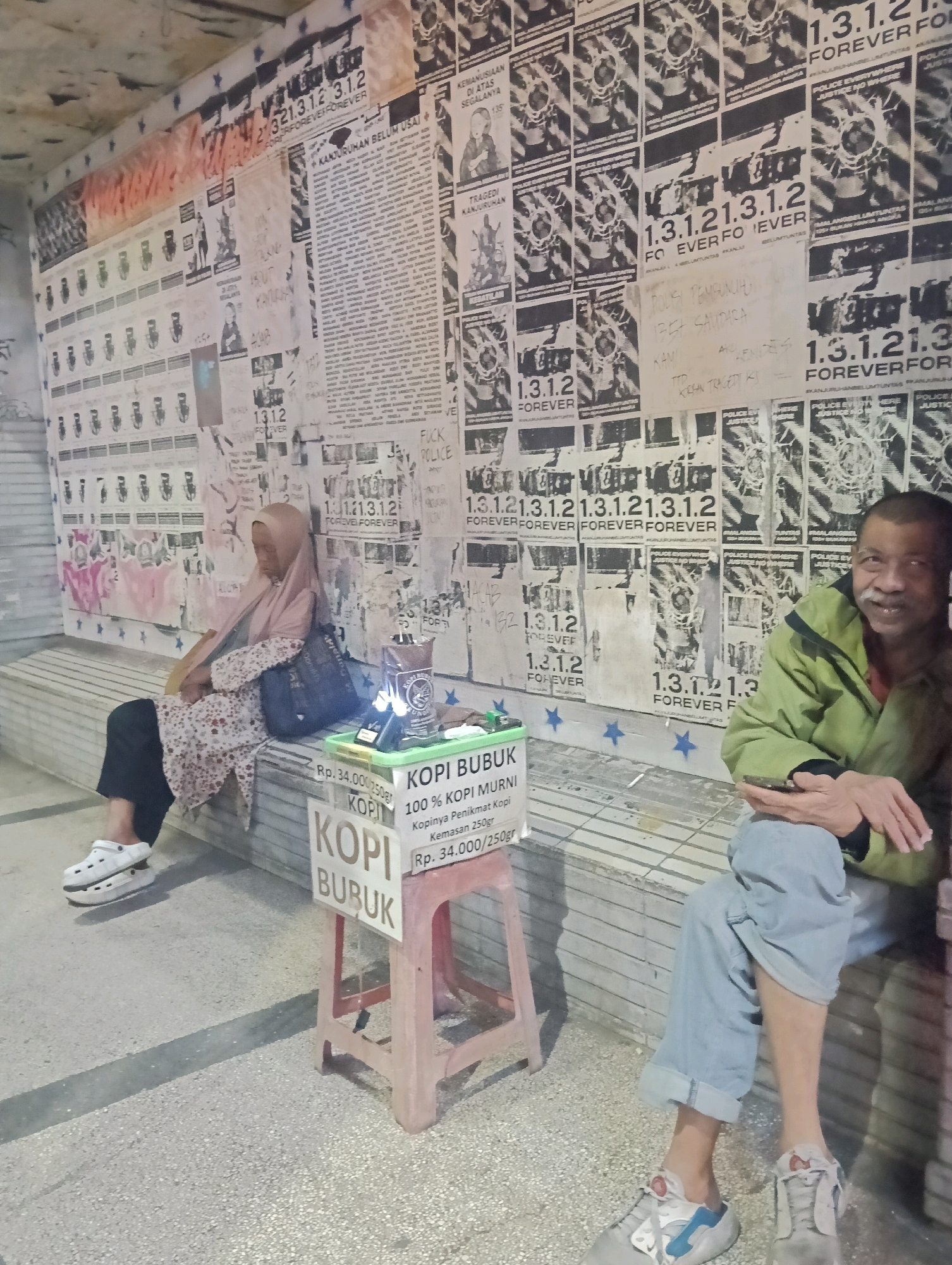 The image depicts an outdoor scene in Malang, Indonesia, featuring a wall covered in numerous posters and graffiti. Two individuals are present, seated on a bench in front of the wall. On the left, a woman wearing a headscarf and a patterned dress is seated, with a dark bag beside her and wearing white platform Crocs. On the right, a man in a green jacket and light blue jeans is seated, holding a mobile phone. In the foreground, a small red stool supports a container of coffee and pricing information, indicating "KOPI MURNI 100% KOPI MURNI" and "Rp. 34.000/250gr." The overall atmosphere suggests a public space, possibly a marketplace or a street corner, with the posters and graffiti hinting at a counter-cultural or artistic expression.