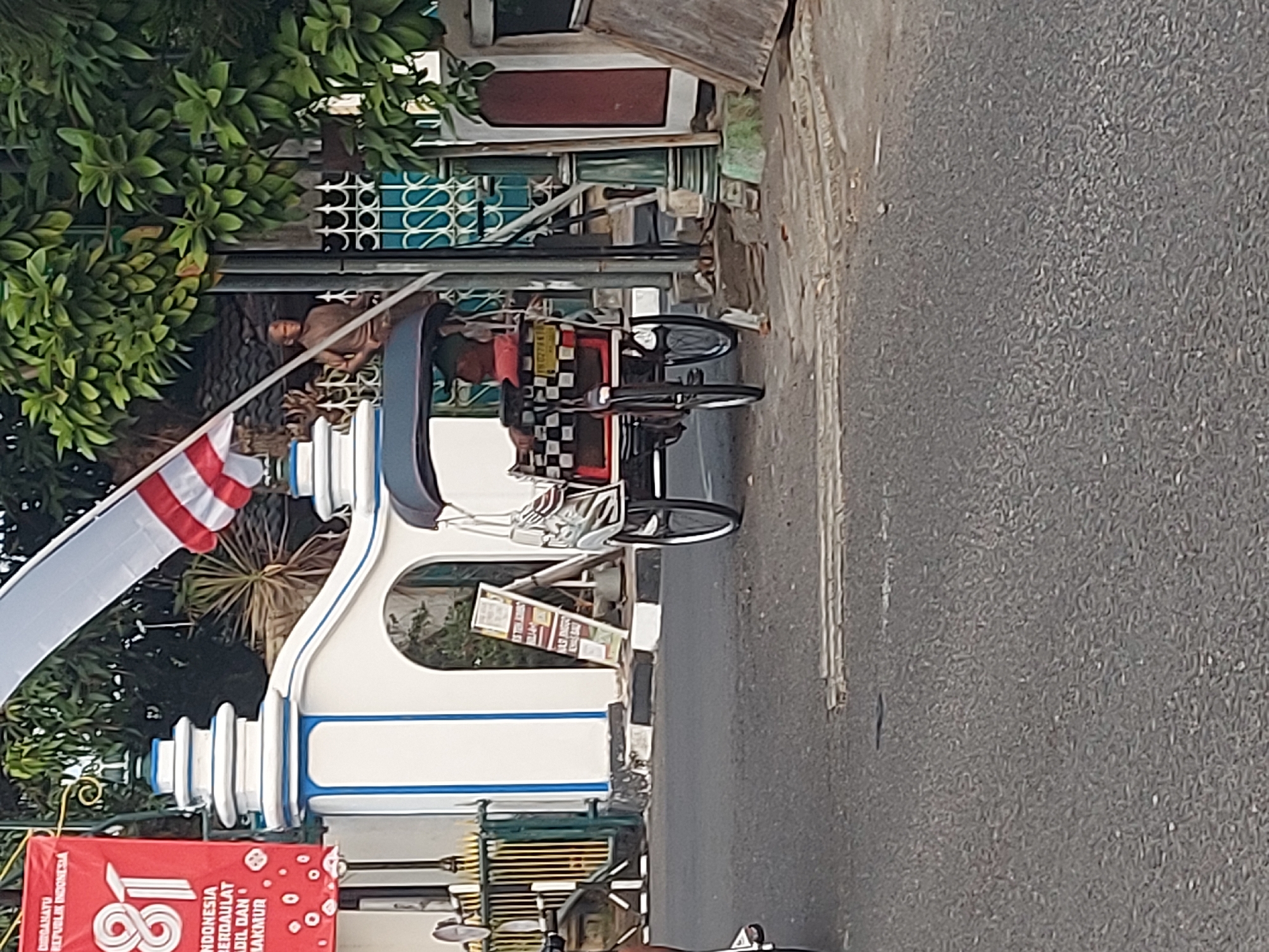 The image depicts a street scene in Yogyakarta, Indonesia, featuring a pedicab parked on the right side of the frame. The pedicab, a three-wheeled passenger vehicle, has a checkered pattern on its rear section and a red cushion. A person is seated inside the pedicab, obscured by the vehicle's structure.
In the background, a white gateway with blue accents frames the entrance to a property. A red banner with white lettering, possibly celebrating Indonesia's 81st Independence Day, is displayed on the left. A statue of a person is visible through the gateway's decorative metalwork. Trees with green foliage are present in the upper right portion of the image. The street itself is paved with asphalt, and a small white marker is visible on the road surface. The overall environment suggests an outdoor urban setting.