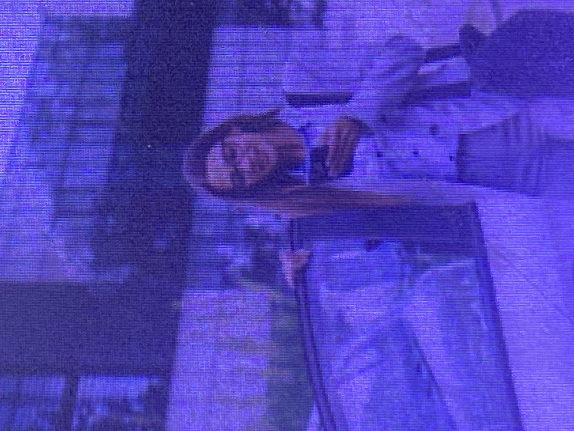 The image shows a person's face reflected in a mirror. The person is looking into the mirror, with their head tilted slightly. Their hair is long and dark, and it appears to be falling over their shoulders. The person is wearing a light-colored shirt. The background is blurry and indistinct, but it appears to be indoors. The lighting is dim and artificial, and the image has a purple cast to it, possibly due to the lighting or a filter. The image is also pixelated, suggesting it may have been taken with a low-resolution camera or has been compressed.
The reflection in the mirror shows a similar view of the person, but with the image reversed. The person's hand is visible, resting on the edge of the mirror. The overall impression is of a candid moment, perhaps a selfie or a photo taken while someone was checking their appearance. The setting is unclear due to the blurriness and the color cast, but it could be a room in a home, a dressing room, or a public restroom. There is no visible text in the image. The location context of Ikorodu, Nigeria, does not provide specific visual cues within the image itself.