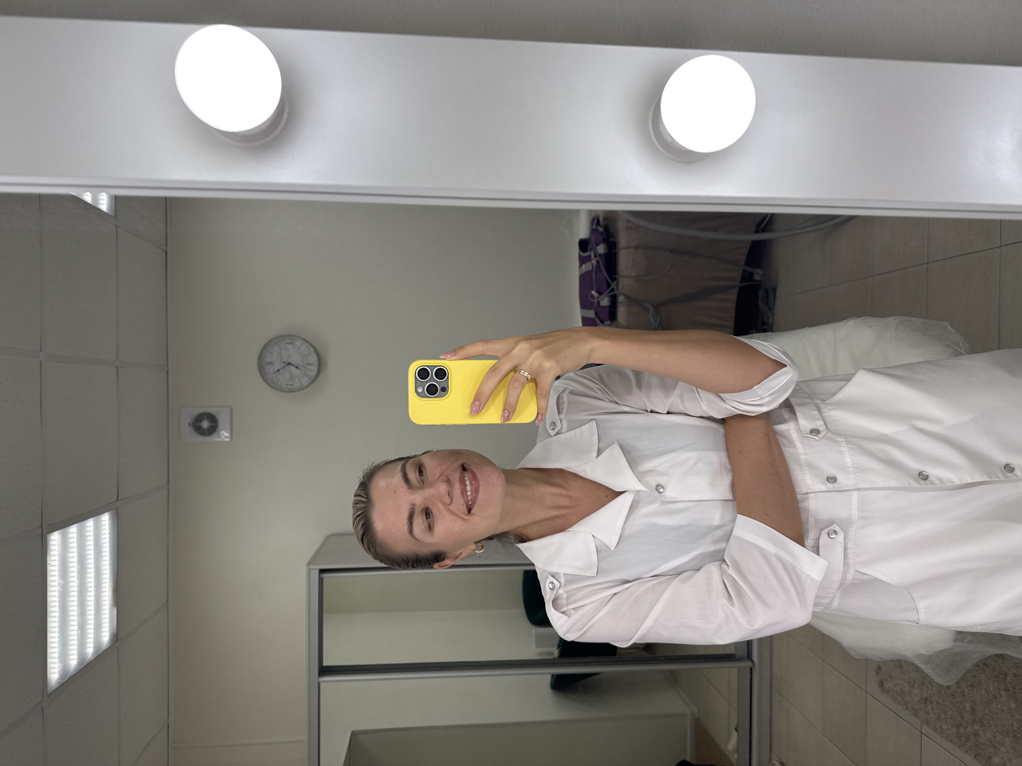 A smiling woman, dressed in a white medical-style uniform with silver snap buttons, takes a mirror selfie. She holds a vibrant yellow smartphone with multiple camera lenses in her right hand, which has light pink manicured nails and a ring on her wedding finger. Her light-colored hair is pulled back, and she looks directly at the phone's camera with a cheerful expression.
The woman is reflected in a large, well-lit mirror, featuring two circular, bright white lights mounted at the top. The background, as seen in the reflection, suggests a professional environment. On a light-colored wall, a round clock with black numbers and hands clearly displays the time as approximately 12:45. Below the clock, a small square ventilation unit is visible. To the left, a ceiling with grid panels incorporates a long, rectangular light fixture emitting bright LED strips.
To the right of the woman's reflection, a white cabinet with glass doors or shelves is partially visible. Further to the right, a white object covered with a disposable sheet, possibly a treatment bed or chair, is present. A purple object, resembling a bag or equipment cover, can also be seen behind her left shoulder in the reflection. The overall lighting is bright and clean, indicating an indoor setting likely a clinic, salon, or professional office. No discernible text is visible other than the numerals on the clock face.