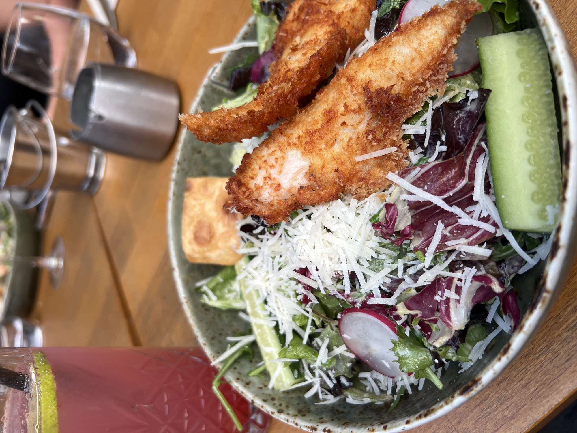 This close-up image showcases a fresh and appetizing salad served in a speckled grey-green bowl on a wooden table, likely outdoors in Plovdiv, Bulgaria. The salad is generously topped with two pieces of golden-brown, crispy fried protein, appearing to be breaded chicken tenders or fish.
The vibrant salad mix includes light green lettuce, reddish-purple radicchio, and is liberally sprinkled with shredded white cheese, possibly Parmesan. Other visible components are thin slices of red radish and a long, thick slice of cucumber. A small, square, golden-brown item, possibly a crouton, is also present in the bowl.
In the lower left foreground, a glass filled with a cold, reddish-pink beverage, complete with a lime slice and a straw, displays condensation. The background is softly blurred but reveals several empty clear glasses and a silver metal container, suggesting a casual dining environment. The natural lighting indicates it is daytime. While no people are clearly visible, the setup implies a relaxed meal or gathering. There is no visible text in the image.