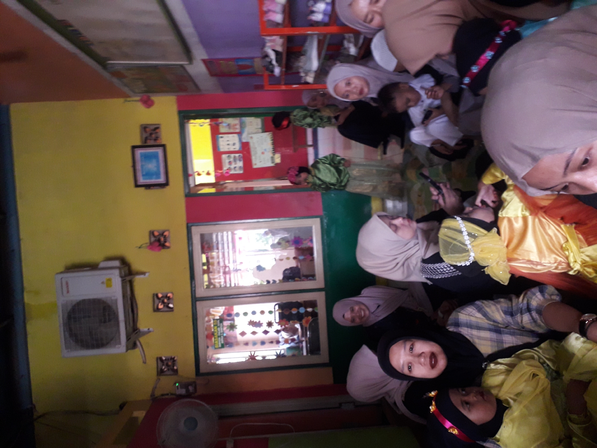 The image shows a gathering of people, mostly women and children, in what appears to be a classroom or community space. The walls are painted in bright colors, yellow and pink, with a purple section on the right. An air conditioning unit is mounted on the yellow wall, and a fan is visible on the left. There are windows with colorful decorations on them. Shelving units with shoes are visible on the right side of the image. Several individuals are wearing hijabs, and some children are in festive attire, possibly for an event or performance. The lighting is somewhat dim, creating shadows. It is implied this scene is in Cankuang, Indonesia.