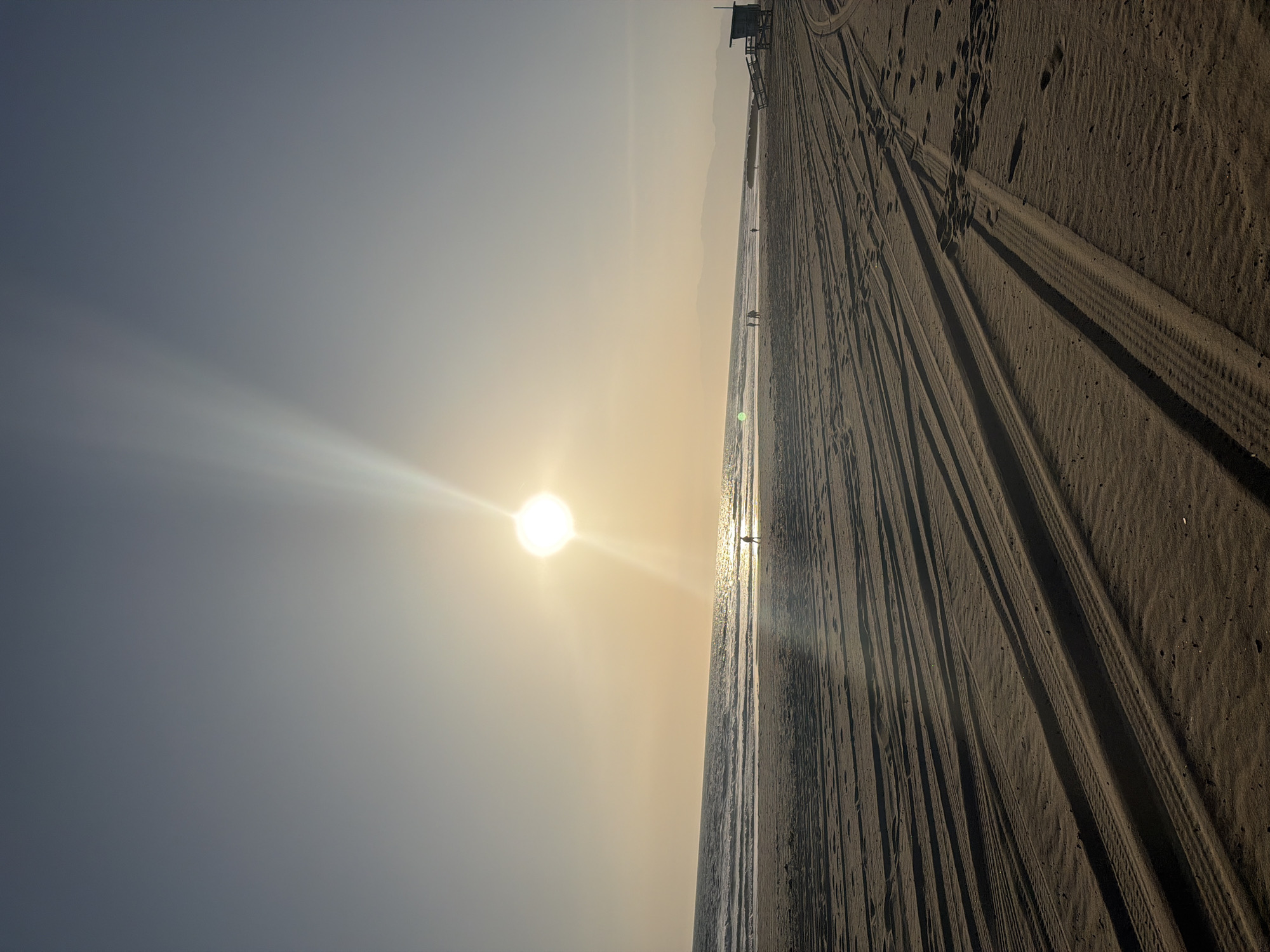 The image shows a beach in Los Angeles, California, at sunset. The sun is low in the sky, casting a bright glare and long shadows. The beach is wide and sandy, with numerous tire tracks. A lifeguard tower is present in the background. Several people are walking along the shoreline. The sky is clear, transitioning from a light blue to a pale yellow near the horizon.