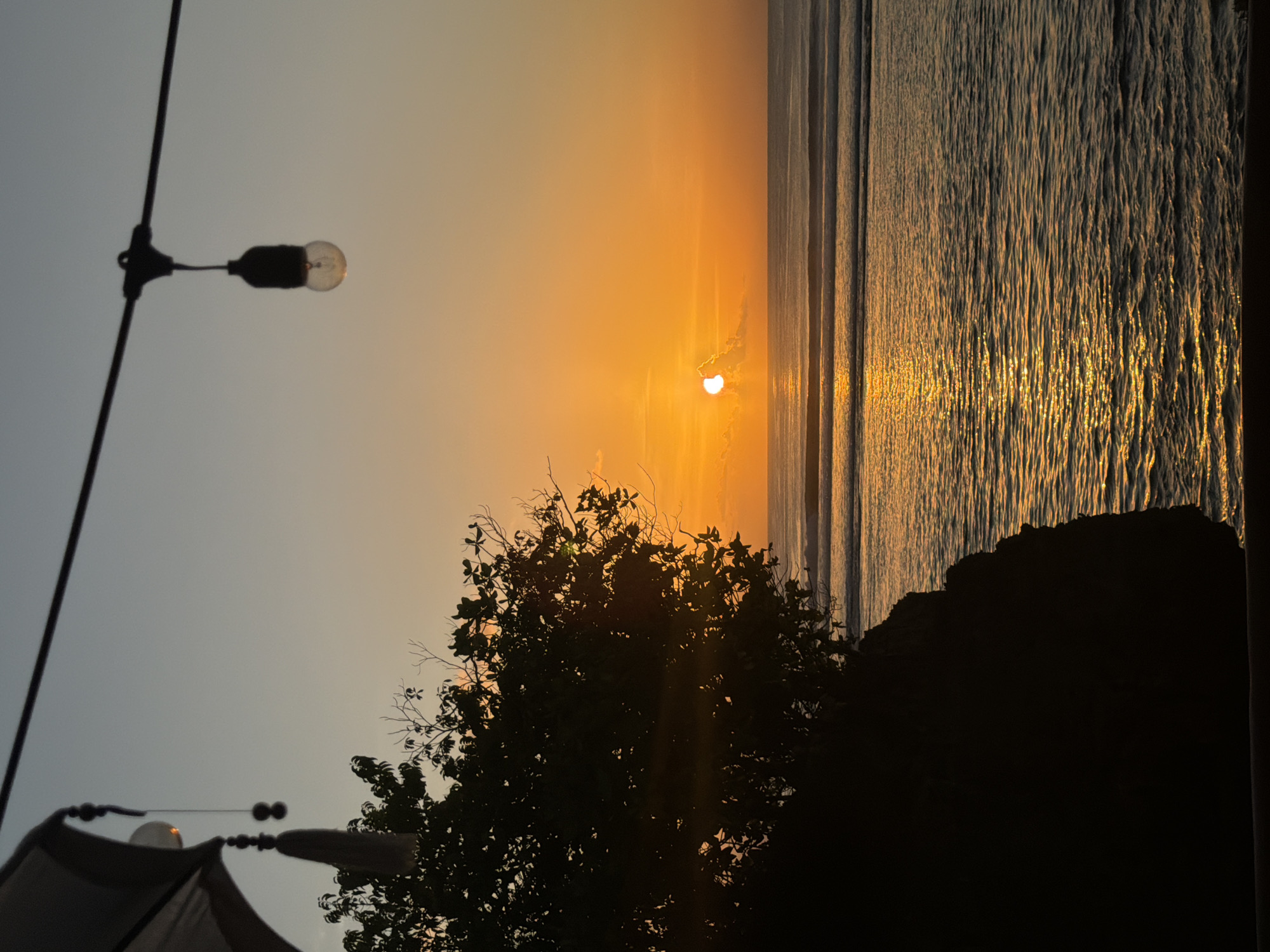 An outdoor scene captures a sunset over the ocean in Kuta Selatan, Indonesia. In the foreground, a silhouette of a rocky cliff and lush foliage obscures the lower left of the frame. A partial view of a white umbrella and decorative tassels hangs from the upper left. A string of outdoor lights is visible across the top of the image, with one bulb illuminated. The sun, partially obscured by a cloud, is a bright orange disc near the horizon, casting a shimmering golden reflection on the water's surface. Several distinct waves are visible on the ocean. The sky transitions from a pale blue at the top to a vibrant orange near the horizon.