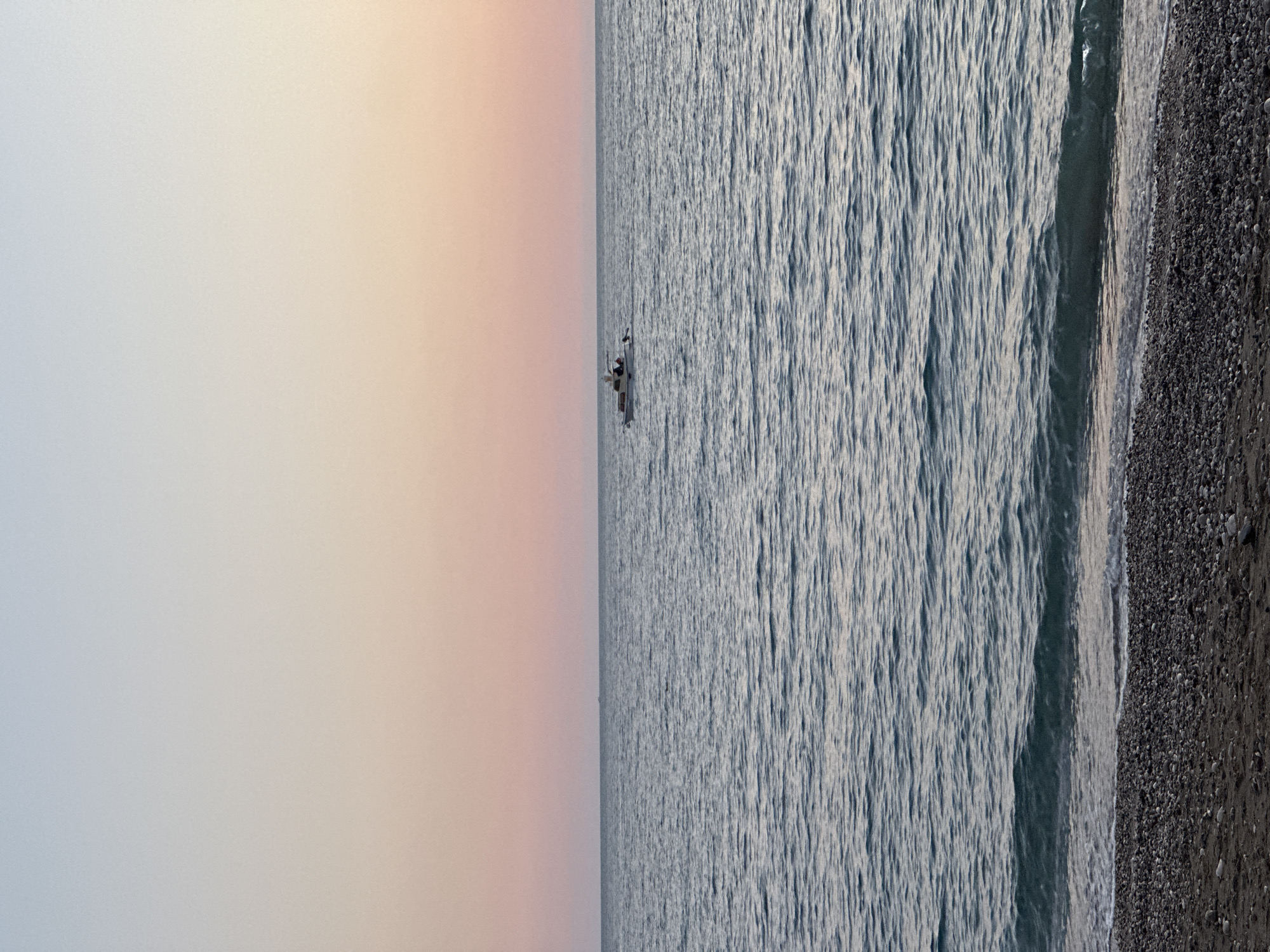 The image shows a horizontal view of the coast of Šušanj, Montenegro. The sky is a gradient of light blue to pale orange. A small boat is centered on the horizon line. The sea is textured with small waves, gradually increasing in size towards the pebble beach on the right side of the frame.