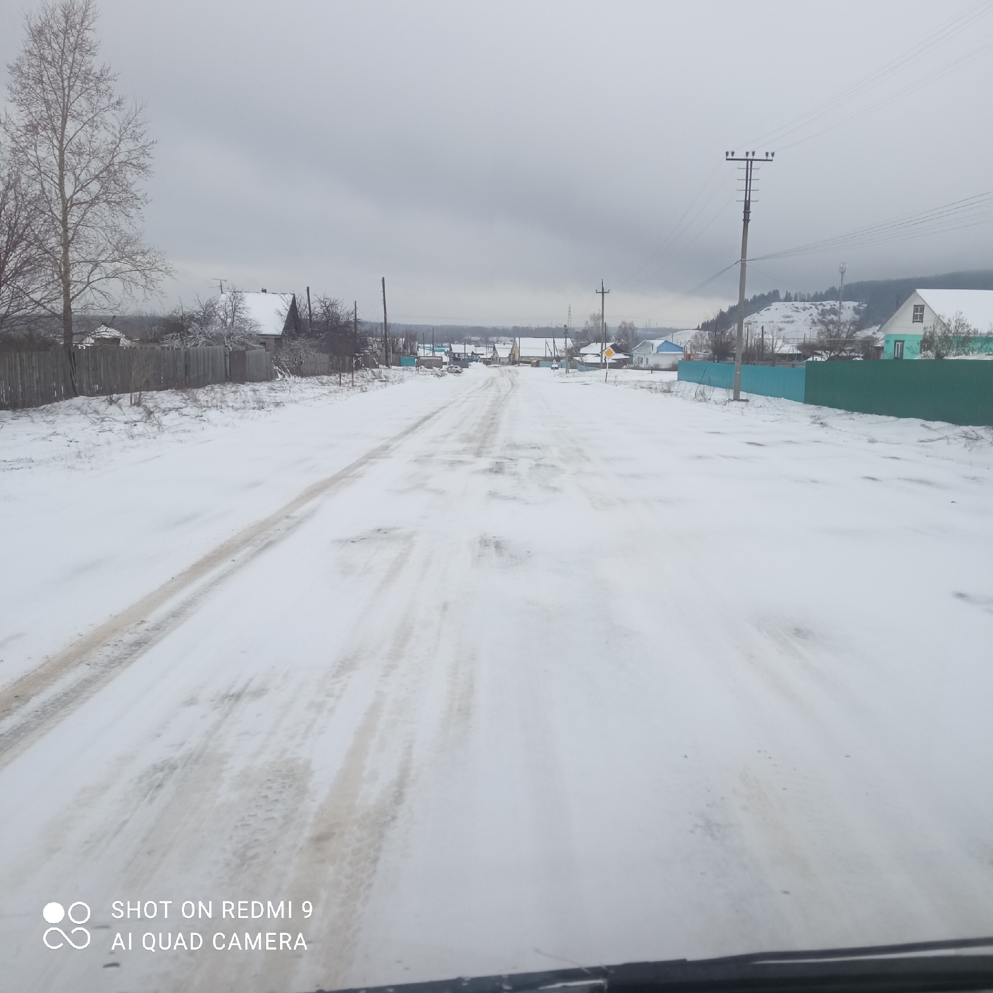 The image shows a snow-covered road in Ольховка, Russia. The road is lined with fences and houses. There are utility poles along the side of the road. The sky is overcast. The photo was taken on a Redmi 9 phone.