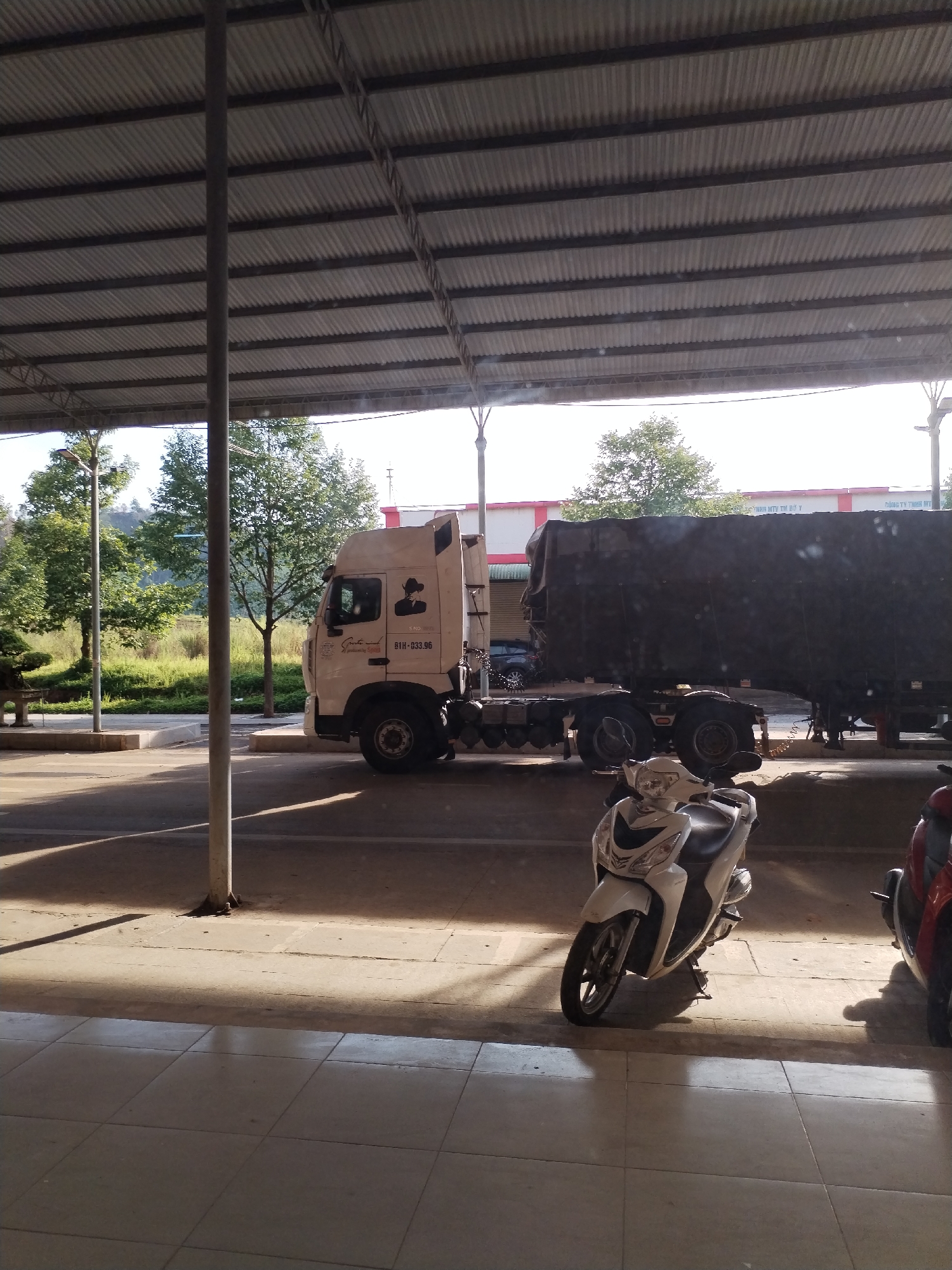 The image depicts a partially covered outdoor area with a corrugated metal roof supported by metal poles, typical of commercial or industrial settings. A white and black semi-truck with a trailer sits parked in the background. The truck cab has a visible graphic on the driver's side. To the right of the truck, a white scooter is parked, casting a shadow. The ground is mostly paved, with some areas showing dirt or possible grass. In the far background, green trees and grassy areas are visible under a bright sky. The image is taken from an elevated position, looking towards the area where the truck and scooter are parked. A portion of a red scooter is visible on the right side of the image. The setting may be the city of Bo Y, Vietnam. The license plate number of the truck is 81H-033.96.