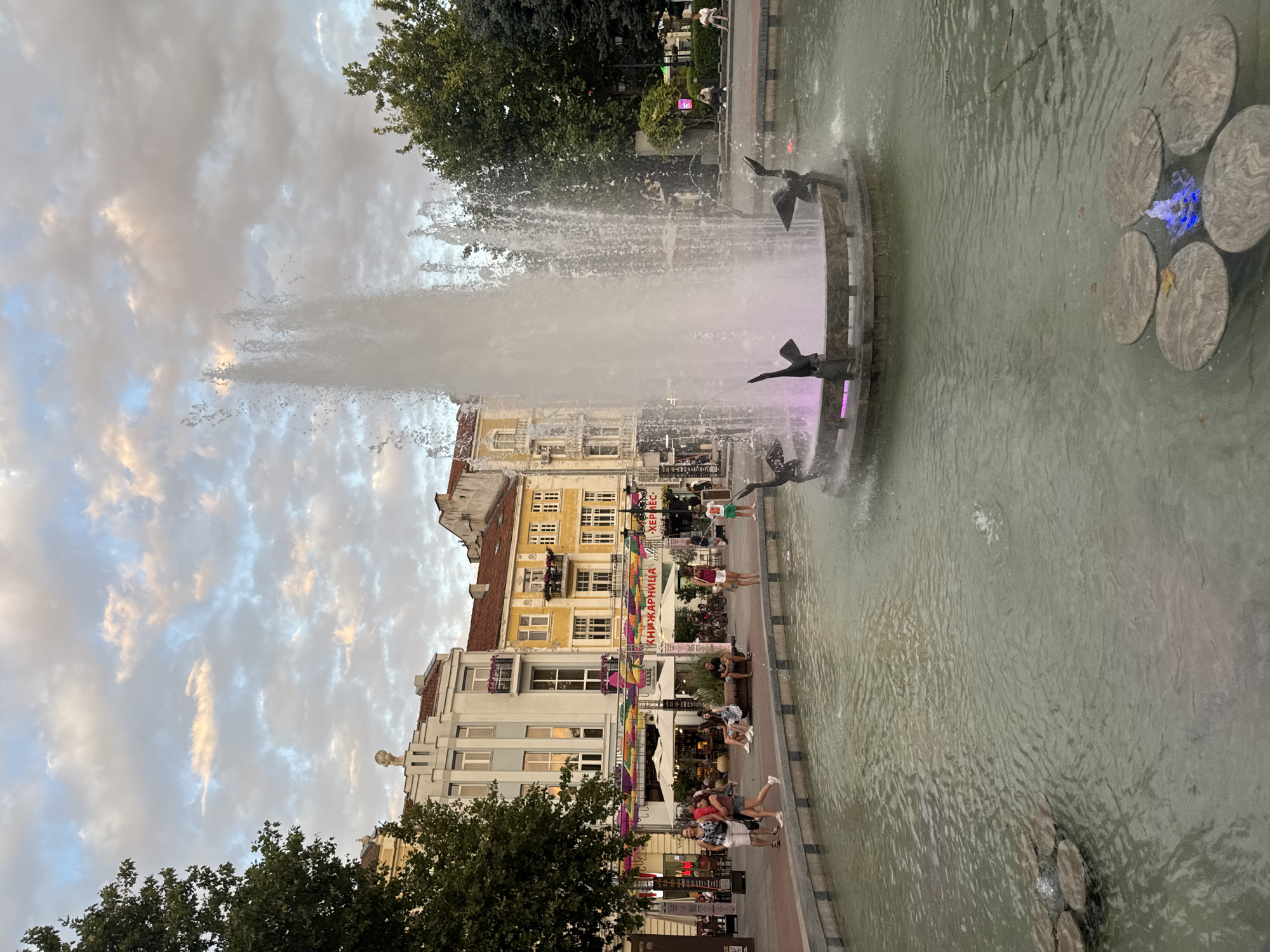 The image depicts a lively city scene in Plovdiv, Bulgaria, likely in the city center, featuring a prominent fountain. The fountain, with its tall spray of water and decorative bird sculptures, is the central element, illuminated by purple lights at its base. In the background, several buildings with distinct architecture, some appearing to be historical, line a pedestrian walkway. These buildings exhibit a mix of styles and colors, with facades in yellow, white, and light blue.
The walkway is bustling with activity. People are seen walking, sitting on benches, and gathering near storefronts. Several individuals are dressed casually, suggesting a warm day. Cafes and shops are visible along the street, with umbrellas providing shade to outdoor seating areas. Colorful banners or decorations hang between the buildings, adding to the festive atmosphere.
The foreground is dominated by the fountain and its surrounding pool. The water in the pool is a greenish-grey color, with some ripple effects from smaller sprays. Several flat, circular stones are submerged in the pool, with one area featuring a small blue light emanating from beneath the water. Lush green trees are present on the left and right sides of the image, framing the urban landscape. The sky above is partly cloudy, with a mix of white and grey clouds, illuminated by soft, diffused light, indicative of either early morning or late afternoon. The overall impression is one of a vibrant public space in a European city, with a blend of historical architecture and modern urban life.