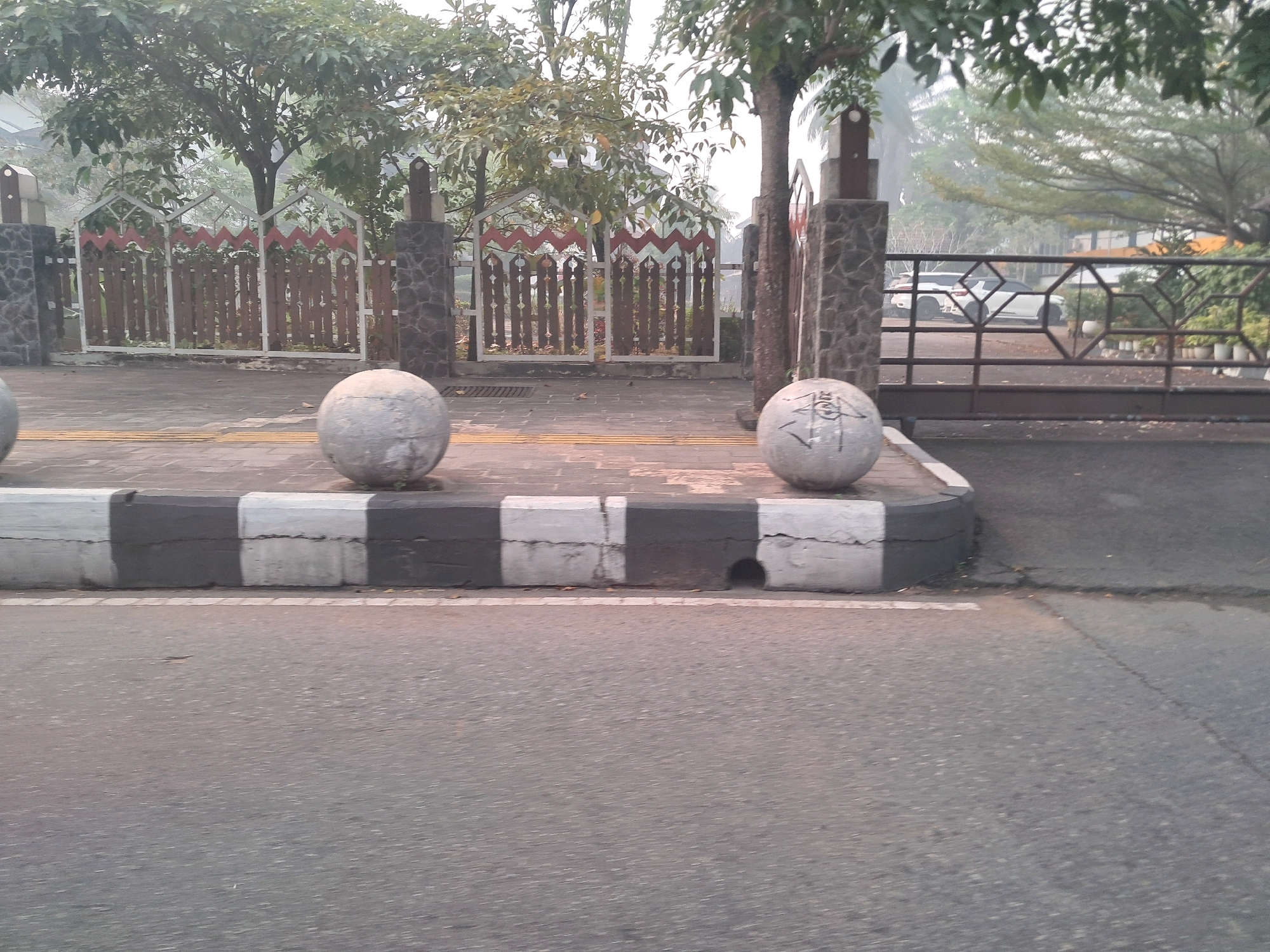 The image shows a street scene in Pontianak, Indonesia, with a gray asphalt road in the foreground. A concrete curb with alternating black and white painted sections runs along the edge of the road. Two large, round, gray concrete spheres are placed on the sidewalk behind the curb. The sphere on the right has some black markings on it. Behind the curb and spheres, a wooden fence with decorative red and white zig-zag patterns is visible, supported by stone pillars. Trees with green foliage are present in the background, some of which are tall and slender. A metal gate with a geometric pattern is also visible on the right side of the image, partially obscuring a white vehicle. The overall atmosphere appears to be somewhat hazy or misty.