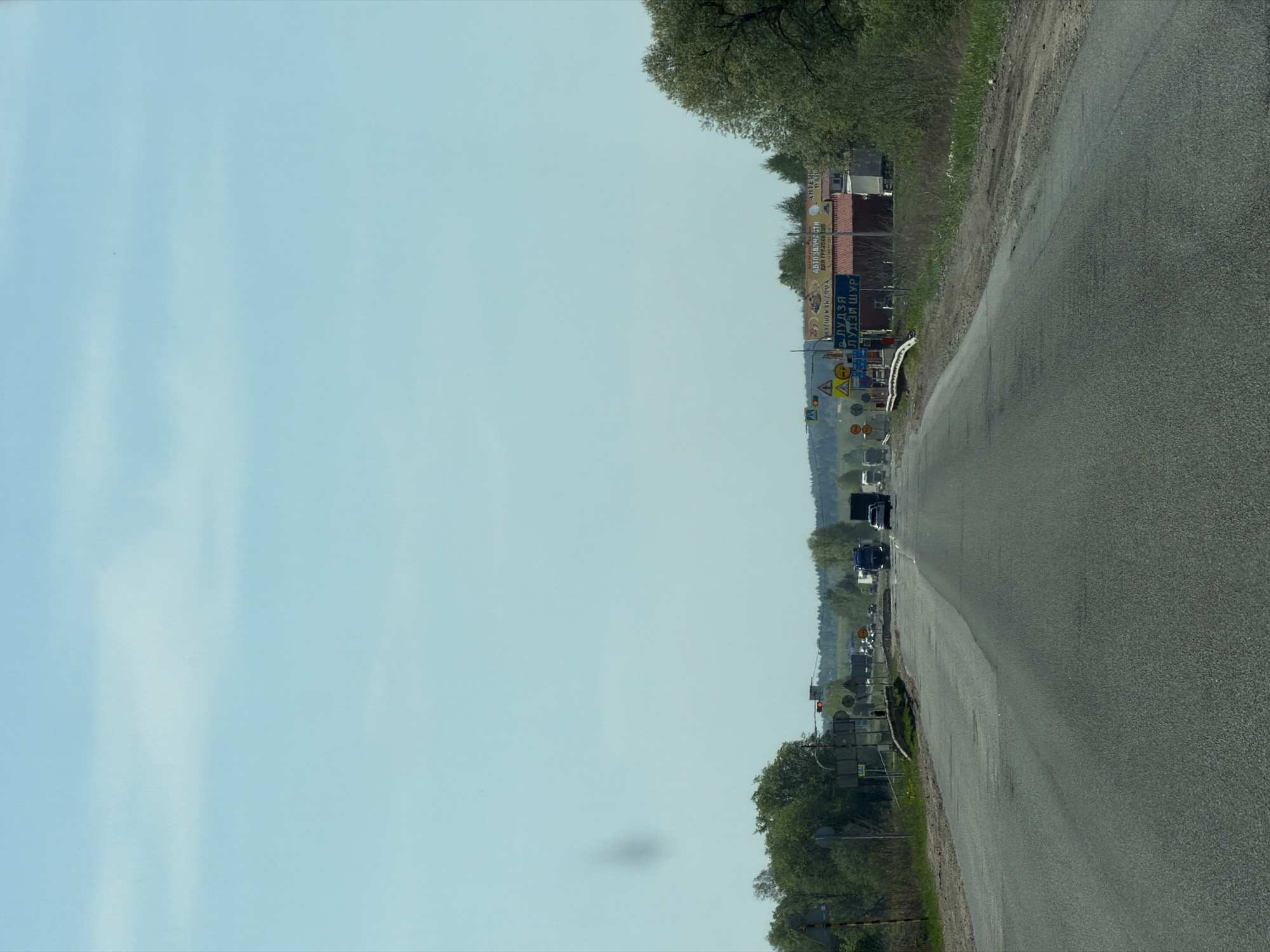 The image shows a road in Совхозный, Russia, with traffic and signage along the roadside. The road surface is asphalt and appears to be slightly weathered. There are several vehicles visible on the road ahead, including what appears to be at least one truck and several cars. A dense collection of road signs and advertising boards are lined up on the right side of the road with Russian lettering. The sky is a light blue and there are green trees and bushes on either side of the road.