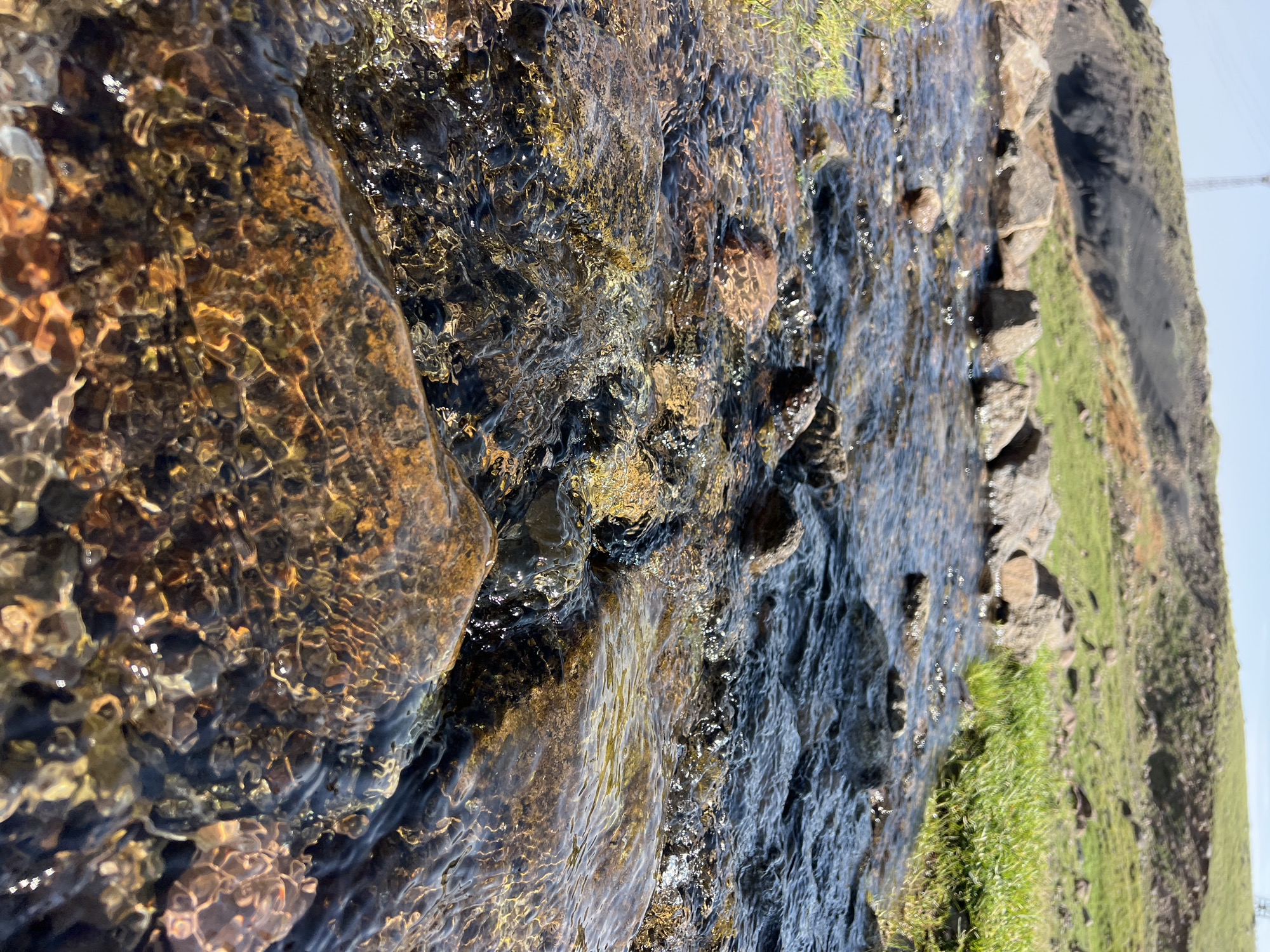 A low-angle, eye-level shot captures a shallow, clear stream flowing over rocks in Aragats, Armenia. The water is dark and reflects the sky, with small ripples and bubbles visible. The rocks in the stream are various shades of brown and orange, with some appearing wet and smooth, while others are rough and textured. To the right of the stream, lush green grass grows along the bank. In the background, a rocky, dark hillside rises steeply, with patches of green grass and some reddish-brown soil visible. A faint, tall metal structure, possibly a utility pole or tower, is visible on the crest of the hill against a clear blue sky. The overall scene depicts a natural, outdoors environment with a focus on the water and its interaction with the rocks.