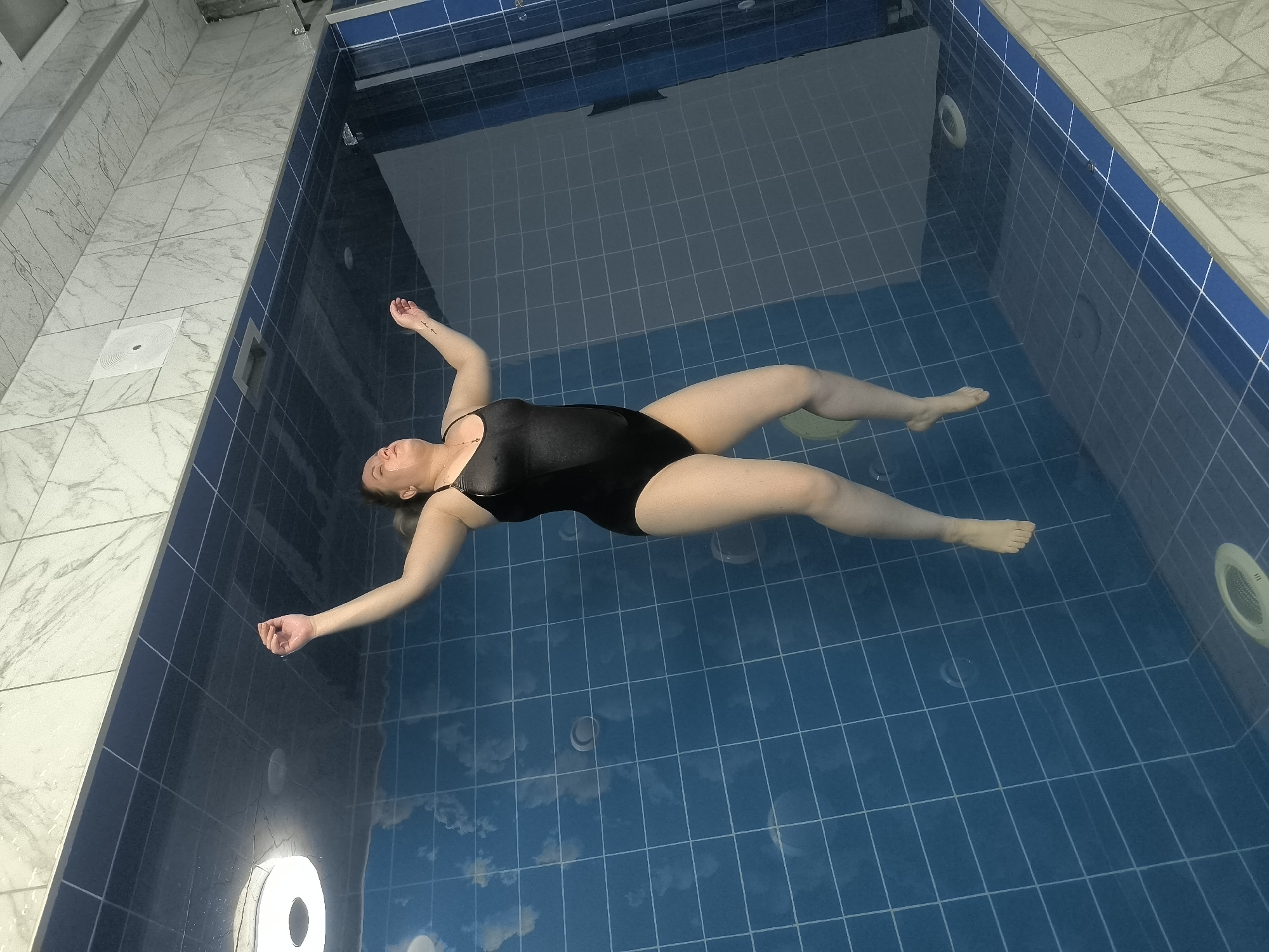 A woman in a black swimsuit floats on her back in a blue-tiled indoor pool. She is positioned diagonally across the frame, with her head towards the upper left and her feet towards the lower right. Her arms are outstretched to her sides, and her legs are slightly spread. The water is clear, and the tiled floor of the pool is visible beneath the surface, marked with a grid pattern. The walls of the pool are also tiled, with a blue border at the waterline. In the background, the tiled floor of the surrounding area is visible, with a white drain cover. Two circular light fixtures are visible along the wall of the pool. The lighting suggests an indoor setting, possibly a spa or a private pool. There is no indication of the time of day or weather conditions. The woman appears relaxed, with her eyes closed and her head tilted back slightly. The overall scene conveys a sense of tranquility and relaxation. The location context indicates this is in Kashino, Russia.