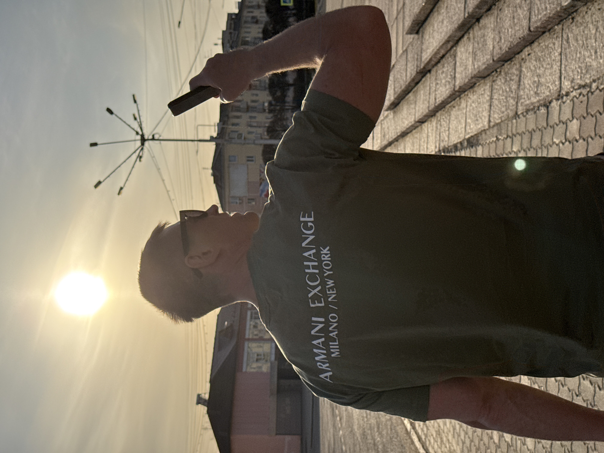 The image shows a person, seen from behind and slightly to the left, standing outdoors in what appears to be an urban setting during late afternoon or early evening. The sun is prominent in the upper left of the frame, casting a strong backlight and creating a lens flare. The person, who appears male, is wearing a dark olive green t-shirt with sunglasses on their head. The text "ARMANI EXCHANGE MILANO / NEW YORK" is clearly visible in white letters on the back of the t-shirt.
The individual is holding a dark rectangular object, likely a smartphone, in their raised right hand, pointing it towards the left side of the frame, possibly taking a photo or video of the sunset or something in that direction. Their head is tilted slightly upwards and to the left, looking towards the bright sun.
In the background, against a clear, bright sky, are silhouettes of buildings and a distinct lamppost with multiple radiating lights. The ground is paved with what looks like patterned tiles or cobblestones. The lighting suggests the "golden hour" with a warm hue to the sky and light. The scene is set in Zelenogorsk, Russia.