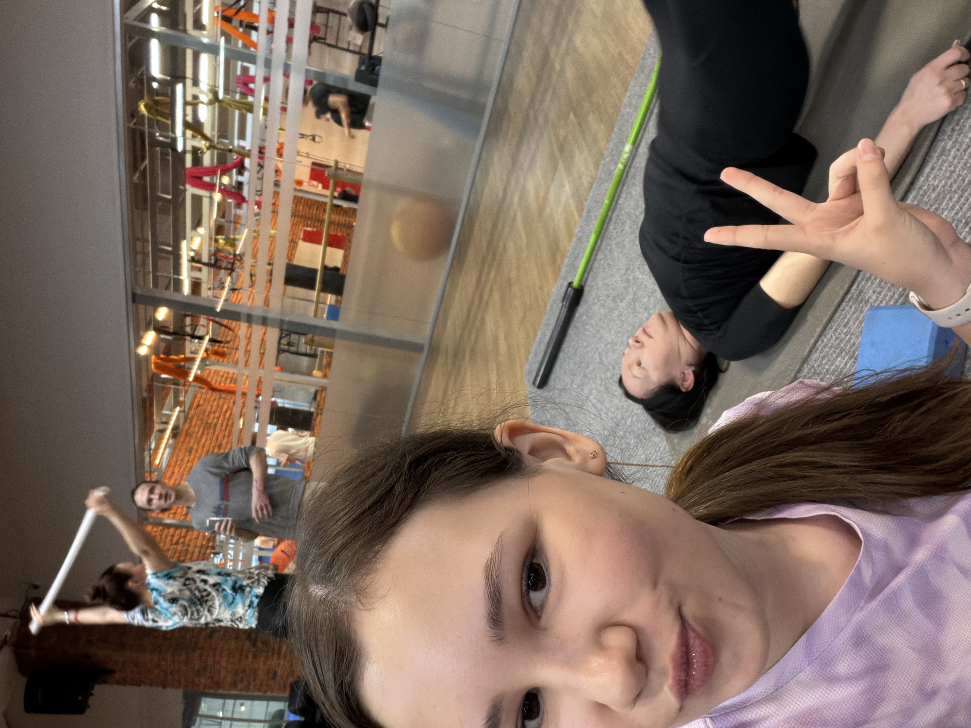 A selfie was taken inside what appears to be a workout facility or studio in the city of Chaikovsky, Russia. A woman is in the foreground, partially in focus, wearing a light purple and white tie-dye shirt. Her dark hair is pulled back from her face. Her right hand is raised, forming a "peace" or "victory" sign.
In the midground, partially in focus, another woman is lying on a gray yoga mat wearing black clothing. A green yoga bar lies next to her.
Behind the first woman, through a glass wall, there is a second workout area with exercise equipment and other people. One man is holding a long white pole or bar and standing next to a woman in a blue and white patterned shirt. Another man in a gray t-shirt is standing further back.