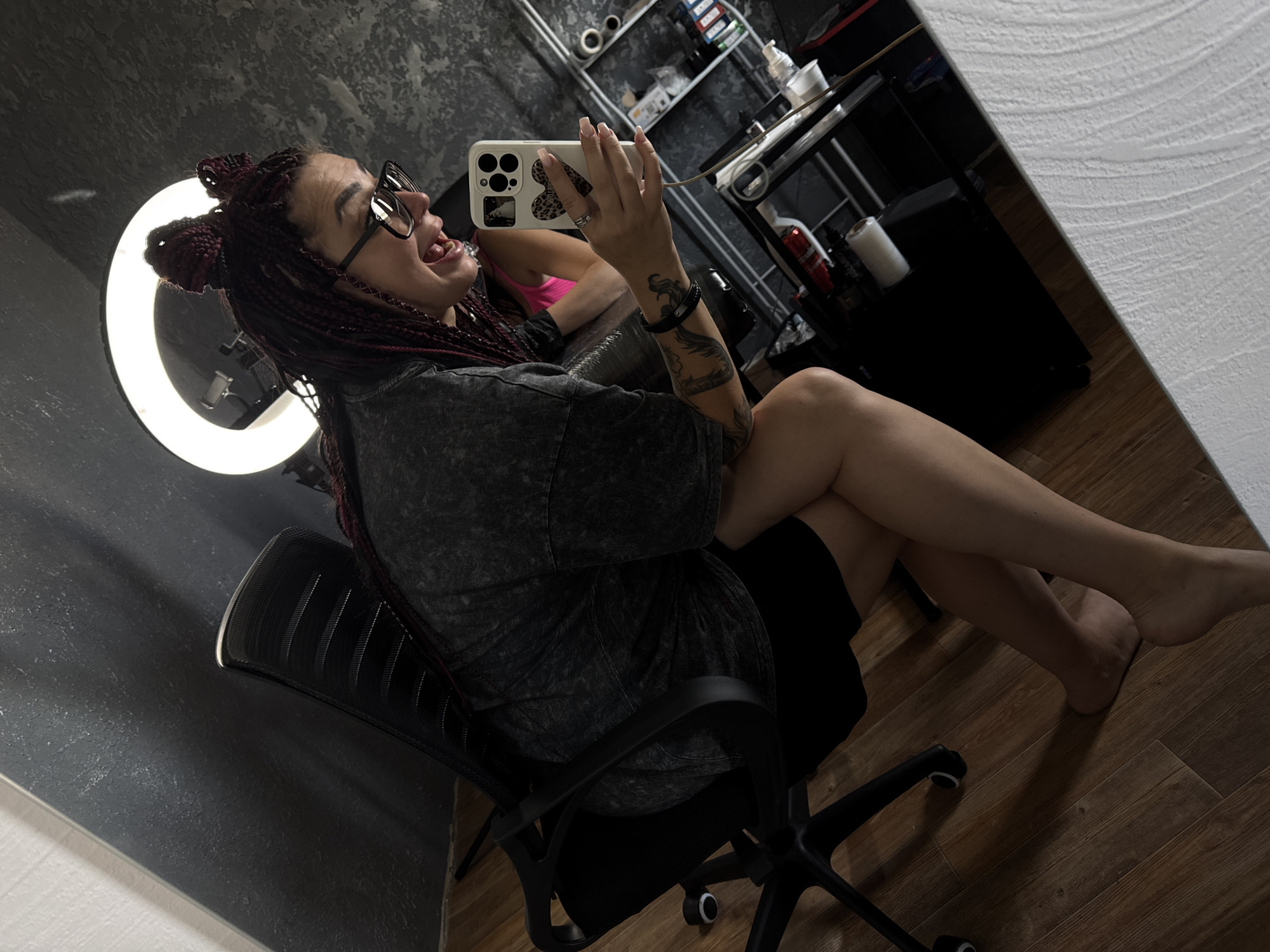 A woman with dark braided hair styled into buns is sitting in a black office chair, taking a selfie with her phone. She is wearing glasses and a dark grey t-shirt. Her tongue is sticking out and her mouth is open, suggesting a playful or excited emotion. Her legs are crossed, and her bare feet are resting on a wooden floor. The background appears to be a room with grey textured walls, a ring light, and shelves filled with various items, possibly in a studio or personal workspace. The overall mood is casual and energetic. No visible text is present in the image.