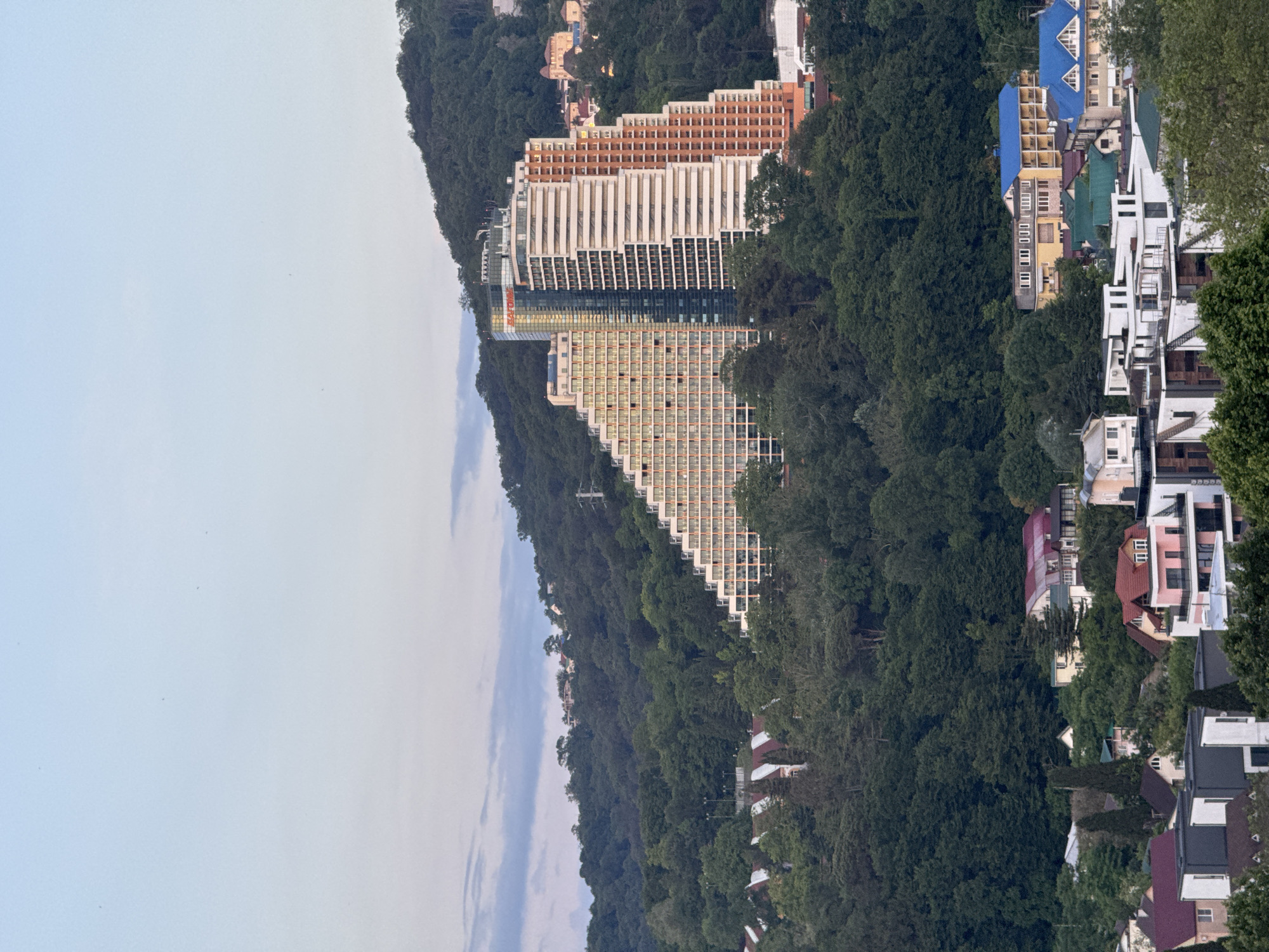 This image captures a wide shot of a city nestled on a densely wooded hillside. In the mid-ground, a prominent, modern hotel complex with a unique tiered design dominates the scene, its light-colored facade contrasting with the dark green trees. The hotel features numerous balconies and windows, arranged in a stepped pattern that follows the contour of the hill. To the left and right of the hotel, more trees obscure parts of the landscape. In the foreground, several residential buildings with colorful roofs, including blue and red, are visible amongst the trees, suggesting a residential area. The sky above is a pale blue with a few wispy clouds, indicating it might be early morning or late afternoon. The overall impression is one of a city integrated with its natural, hilly environment.