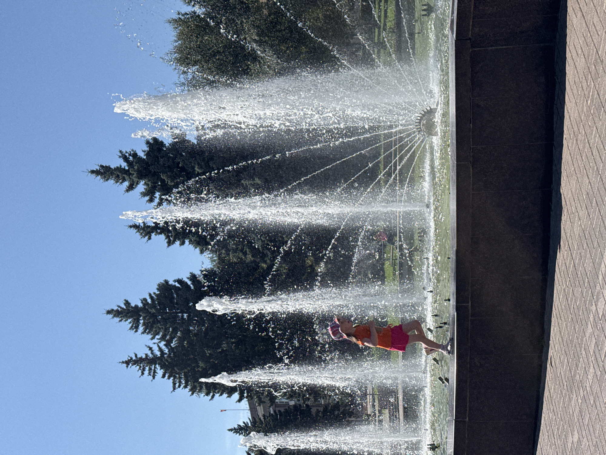 A young girl wearing a pink and orange hat, an orange t-shirt, and pink shorts runs through a fountain on a sunny day. She is wearing sandals. The fountain has several tall water jets spraying into the air. In the background, there are dark green evergreen trees, a building, and a clear blue sky. The fountain is situated on a paved area with a dark stone border. This scene is located in the fountain complex near the "Mir" cinema in Chelyabinsk, Russia.