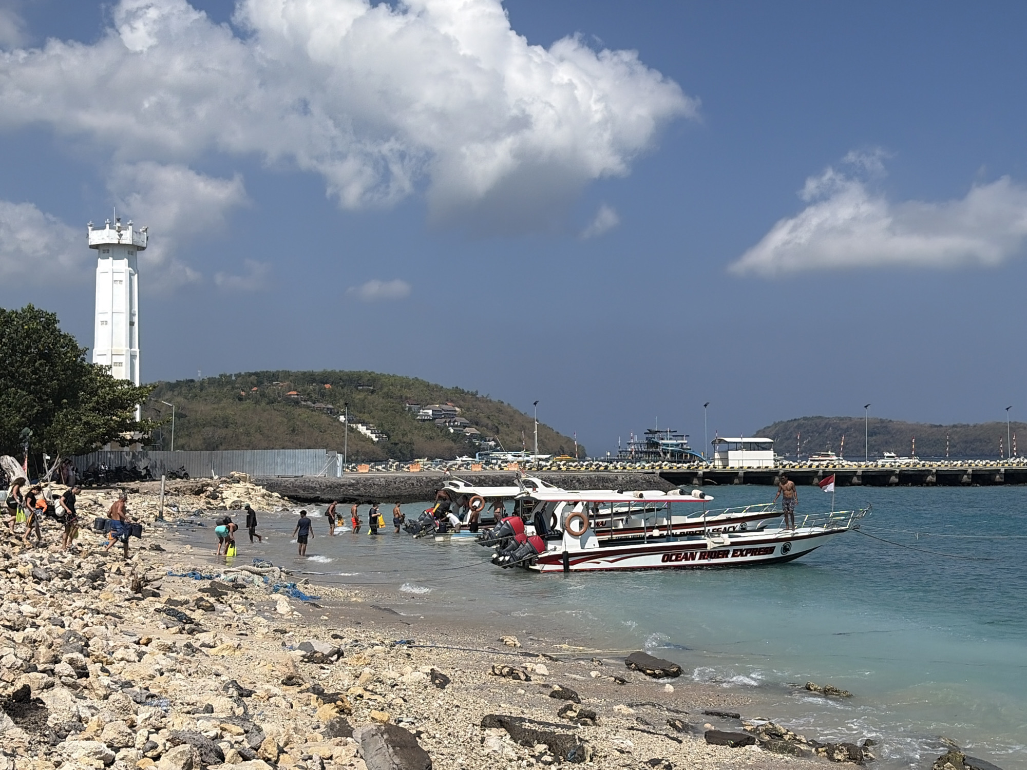 A rocky beach in Nusa Penida, Indonesia, is shown under a bright blue sky with scattered clouds. In the foreground, a rocky shoreline is visible, with some people gathered near the water's edge. Several small boats are moored in the turquoise water, with some appearing to be speedboats. One boat in particular, labeled "OCEAN RIDER EXPRESS," is prominently featured. In the background, a lighthouse-like structure stands tall on the left side of the image. Beyond that, a hillside is dotted with buildings, and a long pier extends into the water, with more boats and structures visible on it. The overall scene depicts a coastal area that serves as a departure point for water activities.