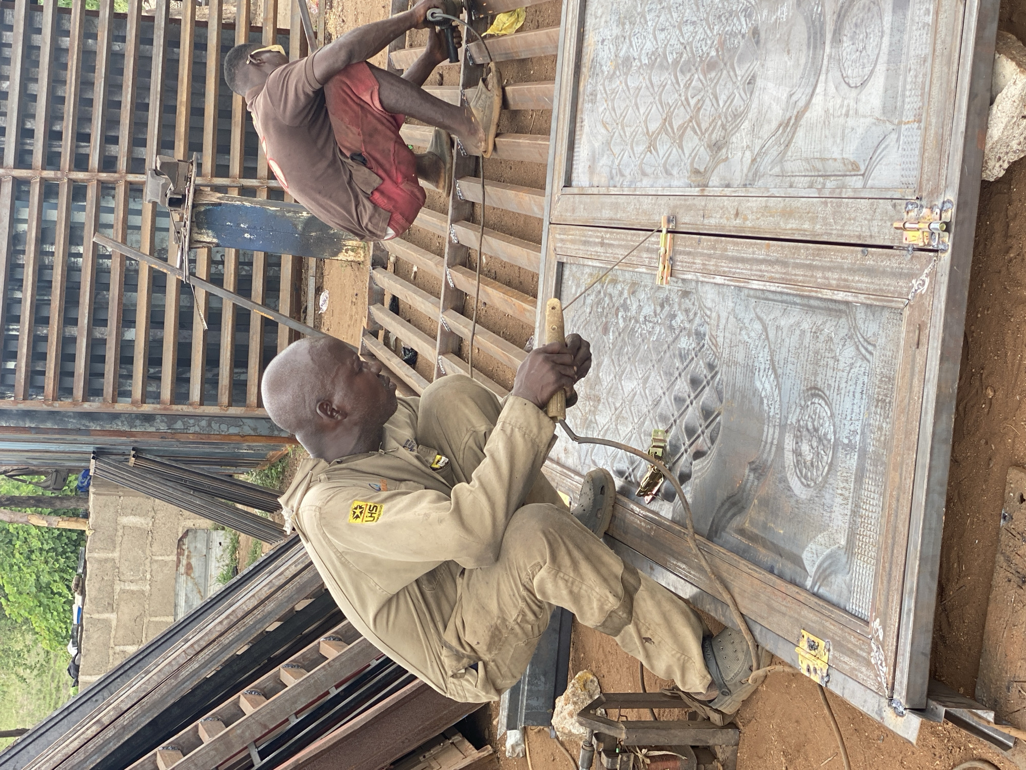 In Adankolo, Nigeria, two men are constructing metalwork outdoors. One man, wearing a brown shirt and red shorts, is perched on a metal structure, operating a grinding tool. The second man, clad in a beige jumpsuit and Crocs, is seated and welding. They appear to be assembling a metal door with decorative panels, secured with yellow latches. Discarded metal pieces and a welding machine are scattered around them on the ground. In the background, there's a stack of metal bars and glimpses of buildings and greenery.