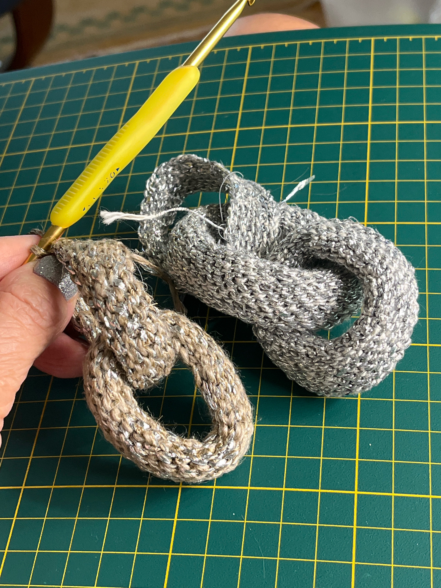 A close-up shot shows a person's hand with a crochet hook in the process of crocheting a loop of metallic yarn in a tan color, which is attached to another, larger crocheted loop of silver metallic yarn. The background is a green cutting mat with yellow grid lines.