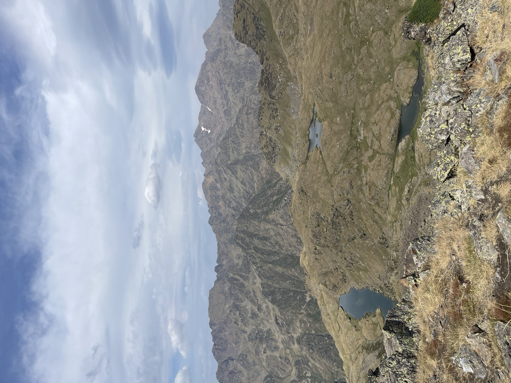 The image depicts a panoramic view of a mountainous landscape under a cloudy sky. In the foreground, rocky terrain with dry grass is visible. Several small, dark lakes are scattered throughout the middle ground, nestled within the undulating, grassy hills. The background is dominated by a range of rugged, brown mountains, some with patches of snow on their peaks. The sky is partly cloudy with a mix of white and grey clouds against a blue backdrop. The overall scene suggests a remote, high-altitude natural environment, likely a mountain range. There are no people or man-made structures visible in the image.