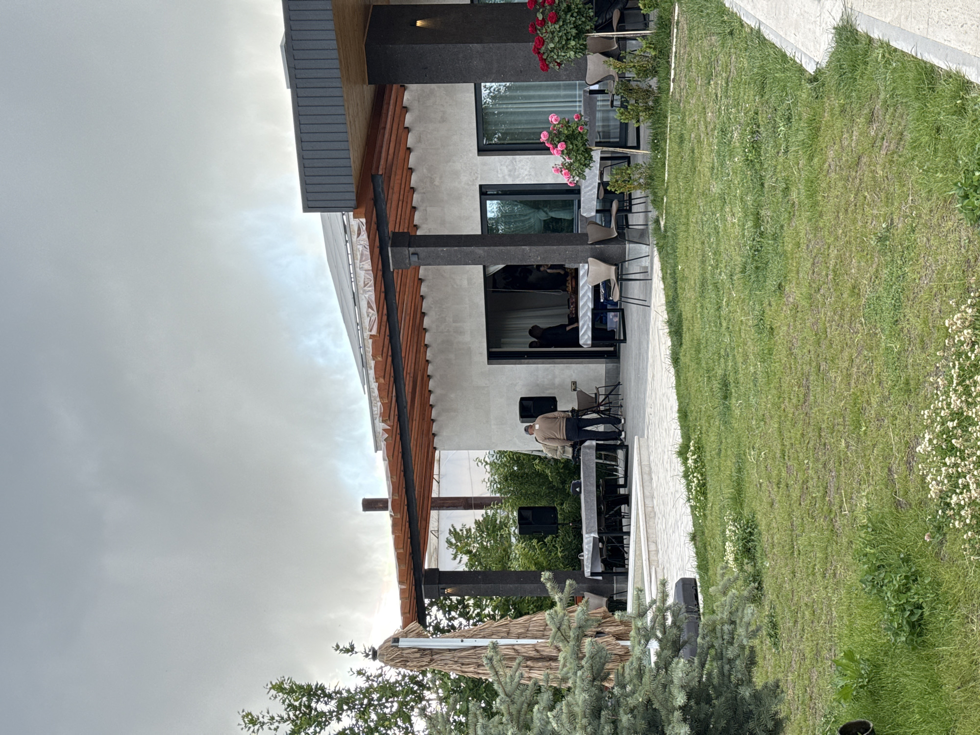 The image depicts the exterior of a building, possibly a restaurant or cafe, in what could be Yerevan, Armenia, judging by architectural style and common features. The structure is a modern design with a light-colored facade, dark support columns, and a wooden-trimmed roof. It features large windows and an open-air patio area with tables and chairs set up. On the patio, there are a few people. One person appears to be setting up sound equipment. There are also potted plants and flowers decorating the facade. The surrounding environment includes a grassy lawn with small white flowers and a paved walkway. The sky is overcast.