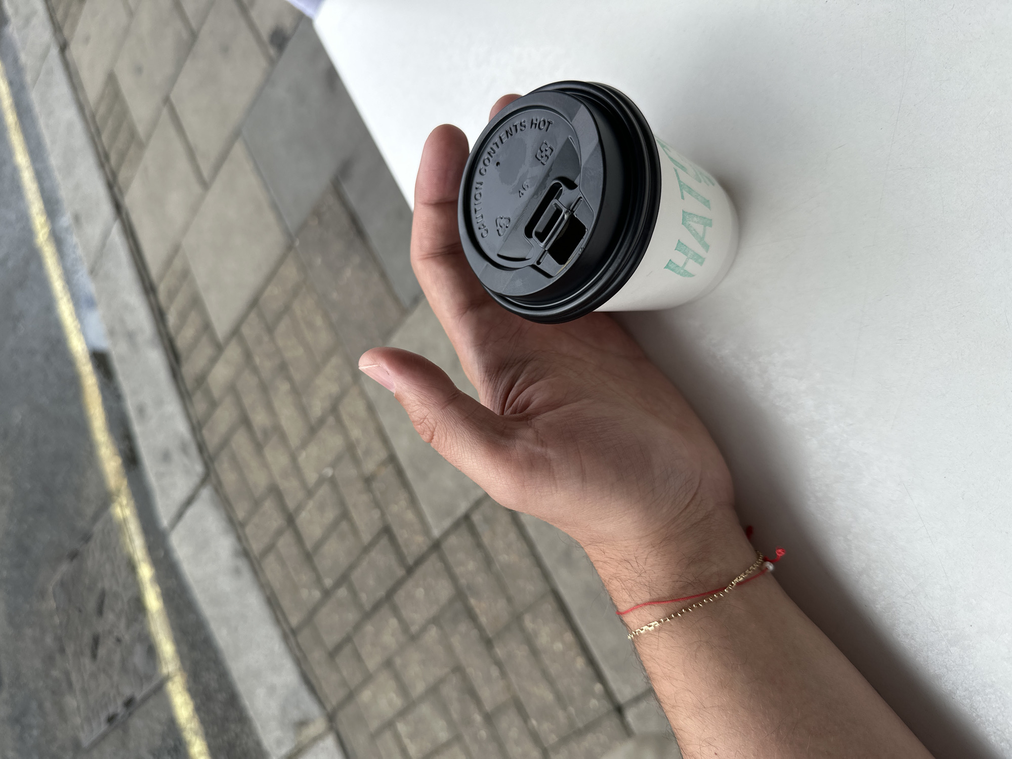 A person's hand holds a disposable coffee cup with a black lid. The cup is white with light green text that appears to read "HATTIS." The lid has "CAUTION CONTENTS HOT" printed on it, along with some symbols and the number "46." The hand has a thin red string bracelet with a small gold chain and a tiny pearl accent.
The background shows two distinct areas. On the left, a textured surface resembling a paved sidewalk with a yellow line running along it is visible, suggesting an outdoor urban environment. On the right, a white, flat surface, likely a table or counter, occupies the rest of the frame.
The image appears to be taken in daylight, possibly late morning or early afternoon, given the lighting. The scene suggests a casual moment, perhaps a person taking a break or on the go, with the coffee cup being the primary focus. The presence of the pavement and the white surface could indicate a cafe or a street setting. No specific emotional cues are discernible from the image. The location context provided is London, United Kingdom.