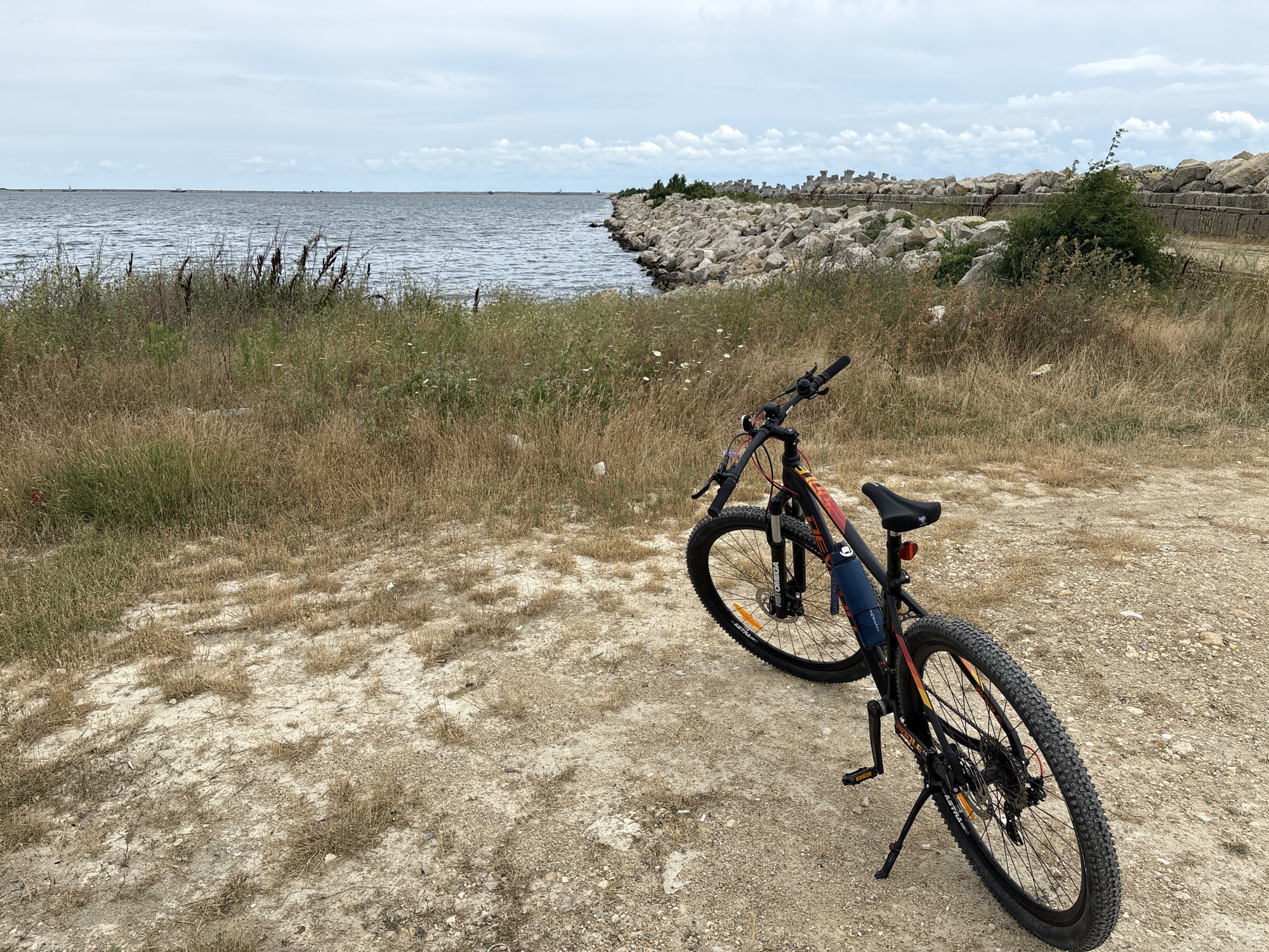A bicycle is parked on a dry, gravelly path next to a body of water. The water is calm with ripples, and beyond it, the horizon is visible under a cloudy sky. Along the edge of the water, there is a rocky embankment made of large stones and some concrete structures. Tall, dry grasses and scattered green vegetation grow along the path and near the water's edge. The bicycle is black with red and yellow accents, featuring thick tires suitable for off-road terrain. It has a kickstand engaged, and a blue water bottle is attached to the frame. The scene appears to be a coastal or lakeside area, possibly within Năvodari, Romania, given the presence of breakwater-like structures.