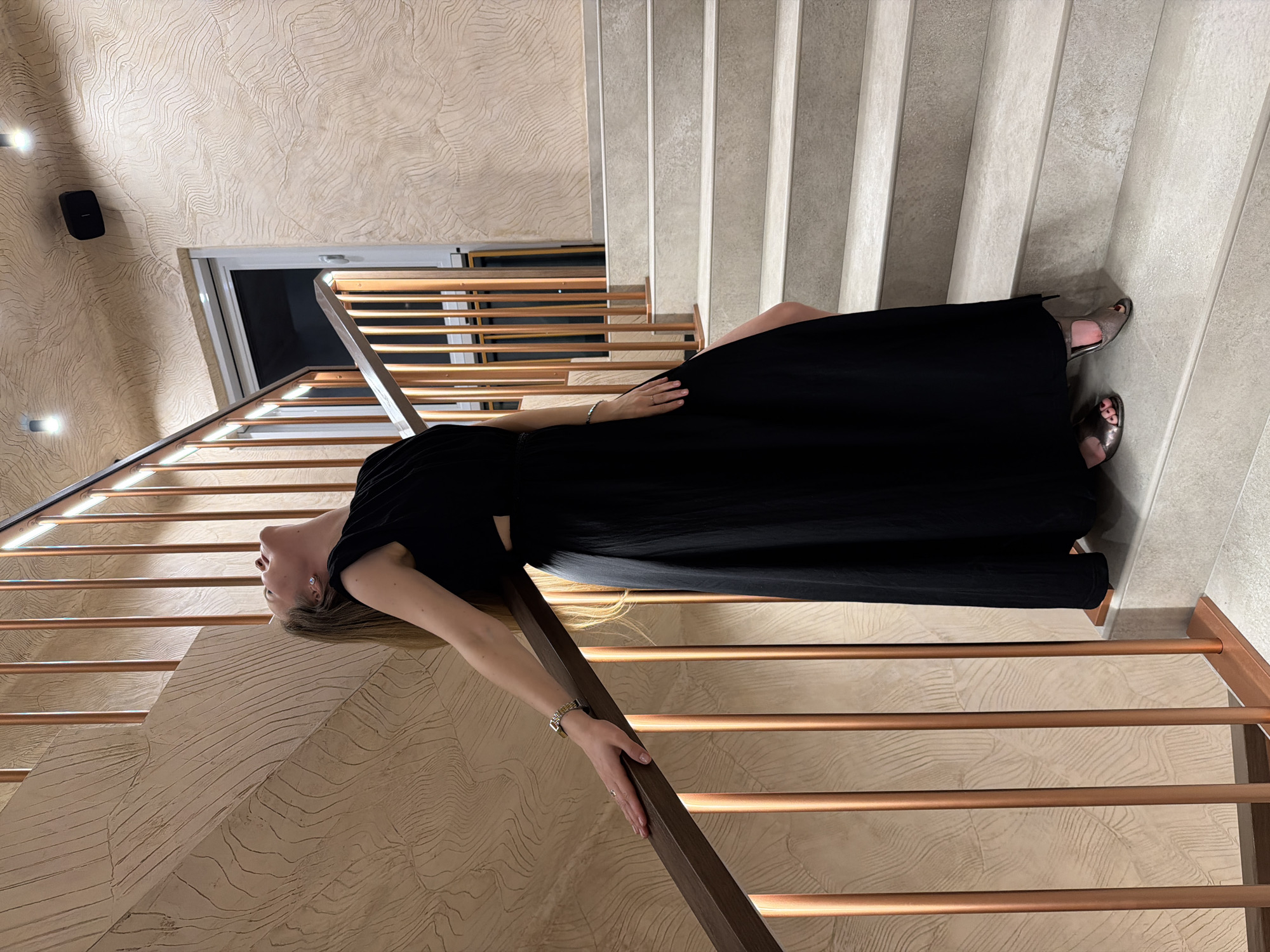 A blonde woman wearing a long black dress and open-toed heels is lying on a staircase. She is holding onto the banister with her left hand. The staircase has beige patterned walls and copper-colored railings. The city of Izhevsk, Russia, is not discernible from the image.