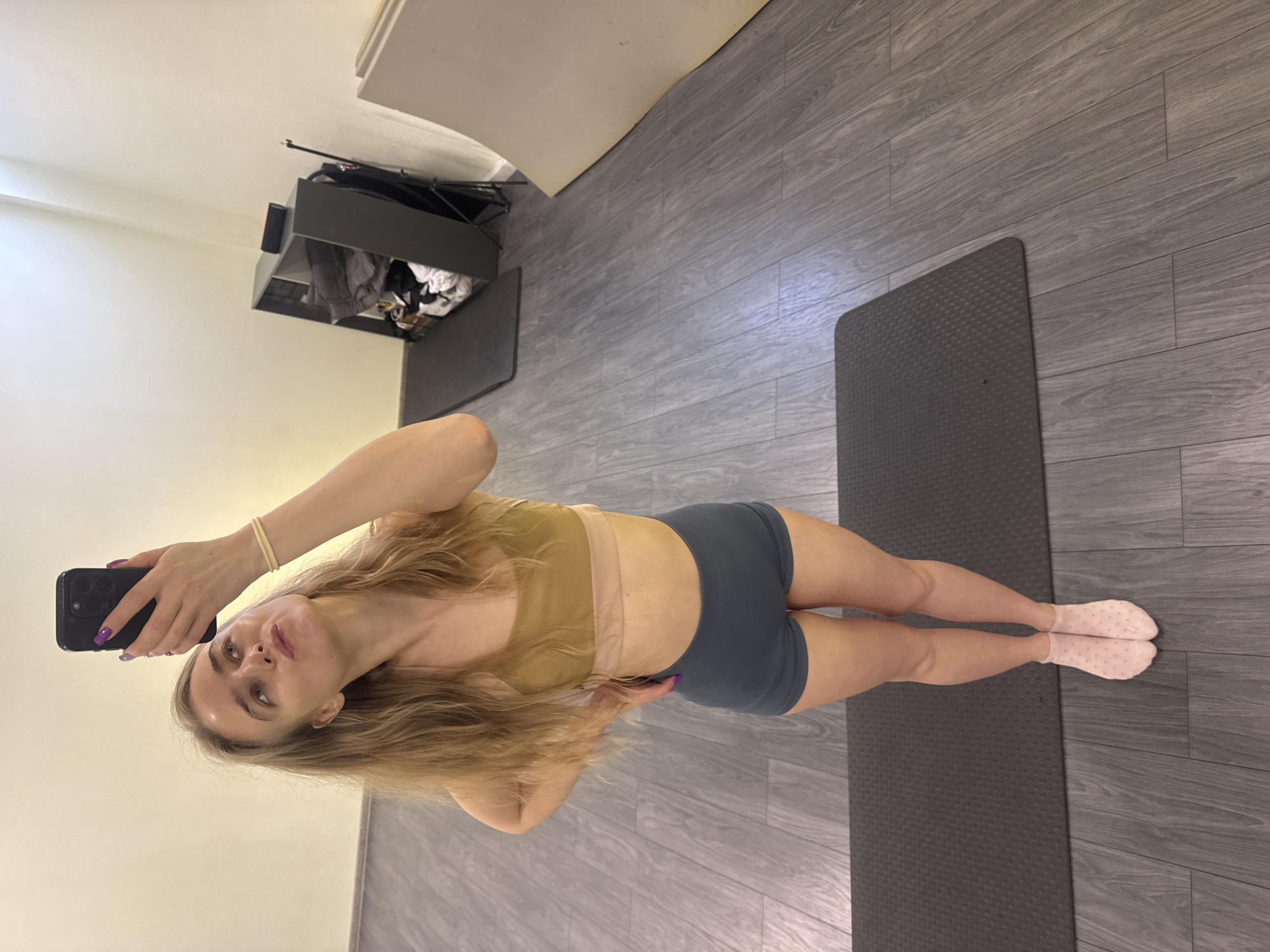 A blonde woman is posing for a selfie in an indoor space with a gray wood-look floor. She is wearing a light brown sports bra and gray shorts with pink socks. A gray yoga mat lies on the floor beside her. In the background is a gray shelf with clothes and other items stored on it, positioned against a white wall.
