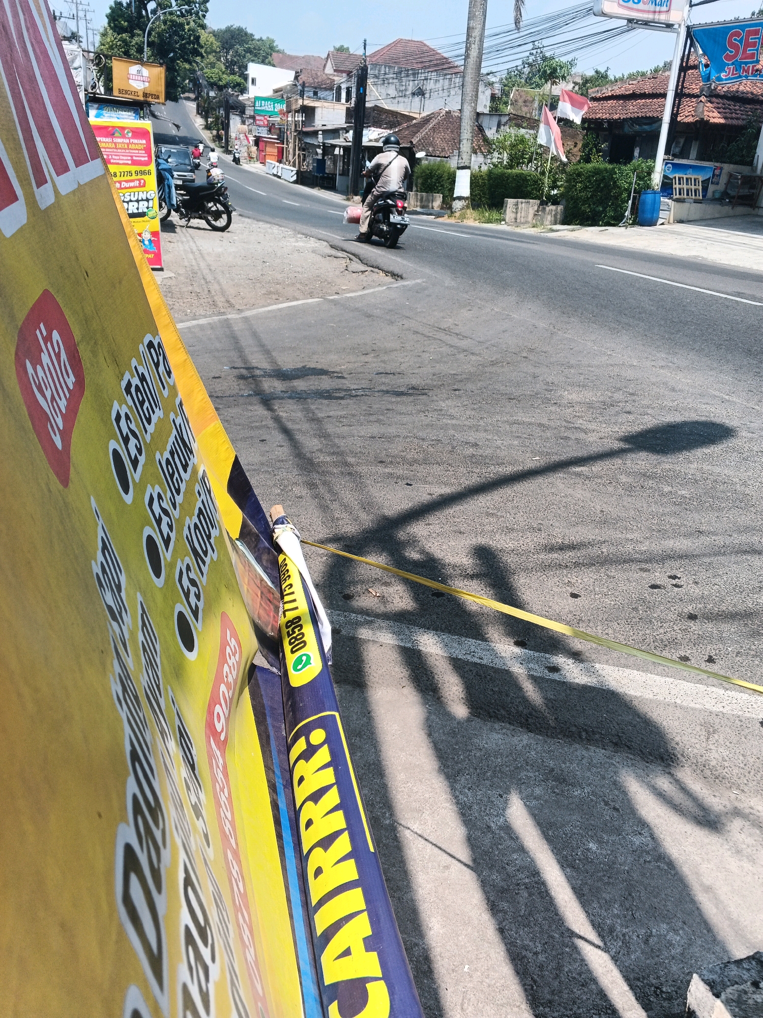 The image depicts a street scene in Semarang, Indonesia, on a sunny day. A yellow and blue banner, partially obscuring the view, displays text in Indonesian, including "Sedia Es Teh / Pa," "Es Jeruk," and "Es Kopi." A phone number "0858 7775 9966" is also visible. In the background, a two-lane asphalt road is visible with a white line marking the lanes. Several buildings line the right side of the road, some with red tile roofs. Two Indonesian flags are displayed, one red and white and the other solid red. A person on a motorcycle is riding away from the camera. Another motorcycle is parked on the left side of the road. A dark-colored car is also parked behind the motorcycle. The shadows of a lamppost and two people are cast on the road. A yellow tape is stretched across the road. The overall impression is a bustling street in a tropical urban environment.