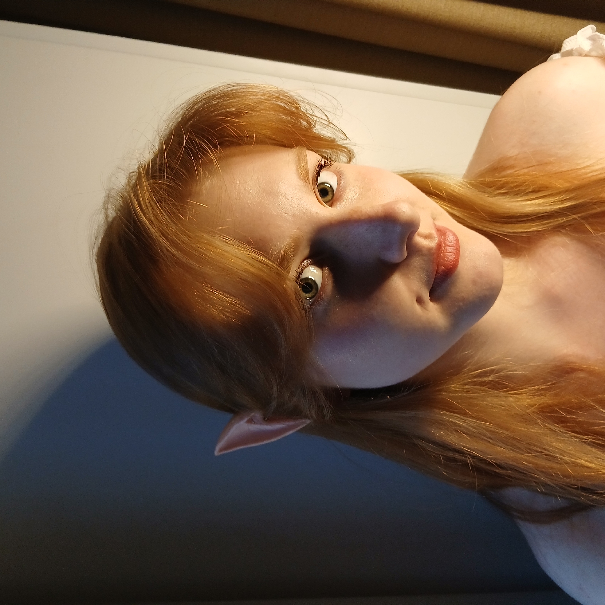 A portrait-style image features a young individual, likely female, with fair skin, bright green eyes, and long, flowing red or auburn hair. The subject is looking directly towards the viewer with an engaging expression and a subtle smile. Their lips are adorned with red lipstick.
A distinctive pointed ear is visible on the left side of their head, suggesting the portrayal of a fantasy character, such as an elf. A hint of a white, ruffled garment can be seen on the upper right, possibly part of a costume.
The lighting is artificial and directional, illuminating the right side of the face and casting the left side and lower hair into soft shadow, highlighting the contours of the face. The background is simple, consisting of a light-colored wall or surface with a darker horizontal element at the top, possibly a shelf or a part of the ceiling.
The image is framed at an unconventional, tilted angle, which adds a dynamic quality to the composition. The overall impression is that of a deliberate pose for a photograph, likely a cosplay or themed photoshoot. There is no visible text in the image.
