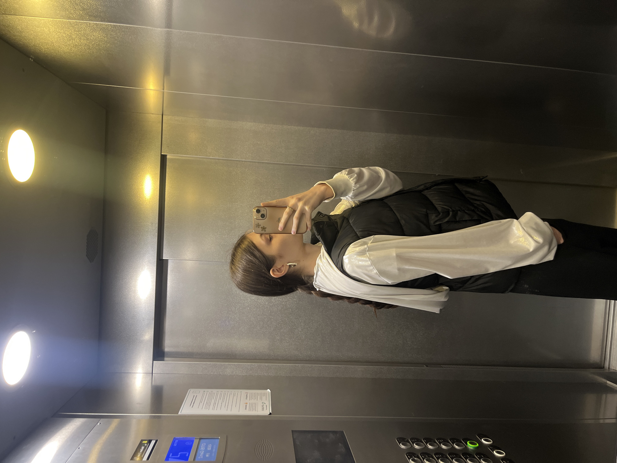 A young woman stands in an elevator, facing a mirror and taking a selfie with her smartphone. She is wearing a white, long-sleeved shirt under a black puffer vest, and black pants. Her hair is pulled back into a braid. The elevator is metallic with a control panel featuring buttons and a small digital display, as well as a printed sign with text. The elevator's lighting is visible as diffused circular lights. The location is not specified in the image.