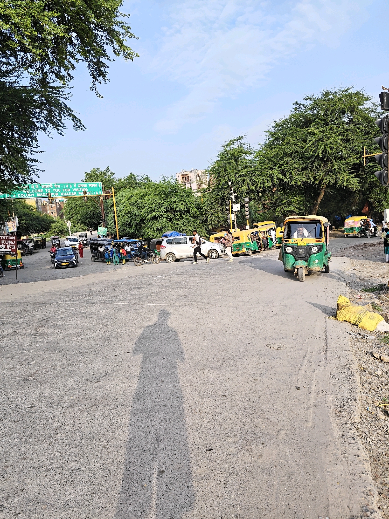 The image shows a street scene in Delhi, India, with a traffic intersection and several auto-rickshaws and cars. In the background, there is a green archway with Hindi and English text that reads "Welcome to You For Visiting J.J. Colony Madarpur Khadar Phase-I & II". The sky is clear with a few clouds. The road is paved and appears to be in moderate condition. Several people are visible, including drivers of the auto-rickshaws and pedestrians crossing the street. A person's shadow is cast on the road in the foreground. There is also a yellow bag on the side of the road.