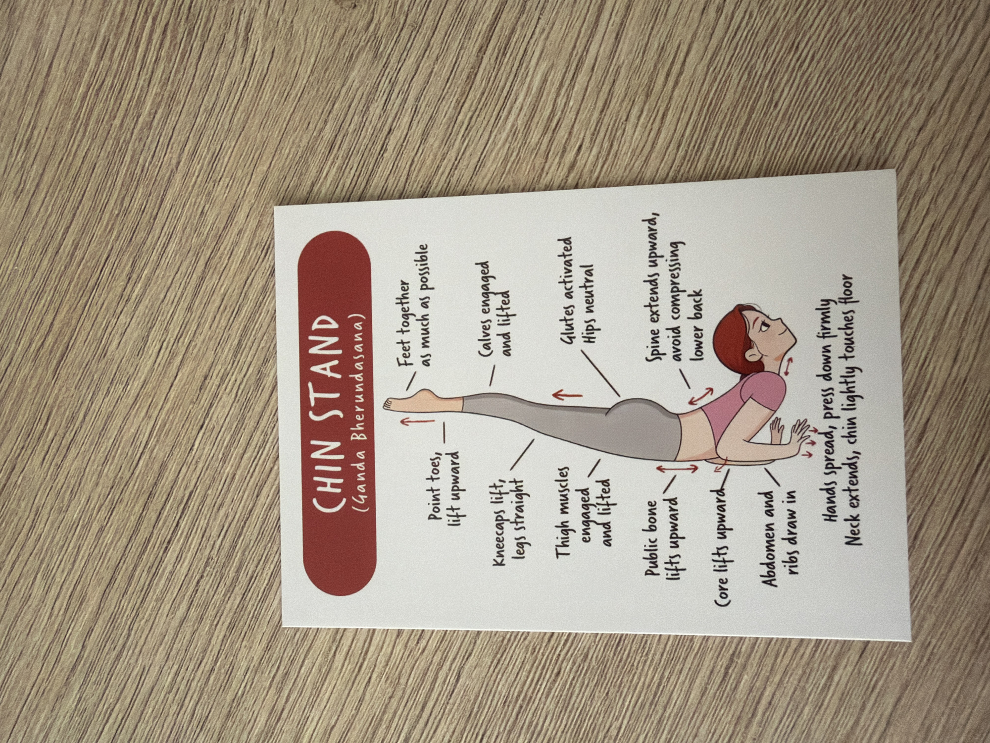The image shows a printed card with instructions for a yoga pose called "Chin Stand (Ganda Bherundasana)". The card features a diagram of a person performing the pose, with various body parts labeled with instructions like "Point toes, lift upward", "Kneecaps lift, legs straight", and "Glutes activated, hips neutral". The card is placed on a wooden surface, which appears to be a table or floor with a wood grain pattern. The lighting suggests an indoor environment. There is no information in the image that would allow for the determination of its location in Pazardzhik, Bulgaria.