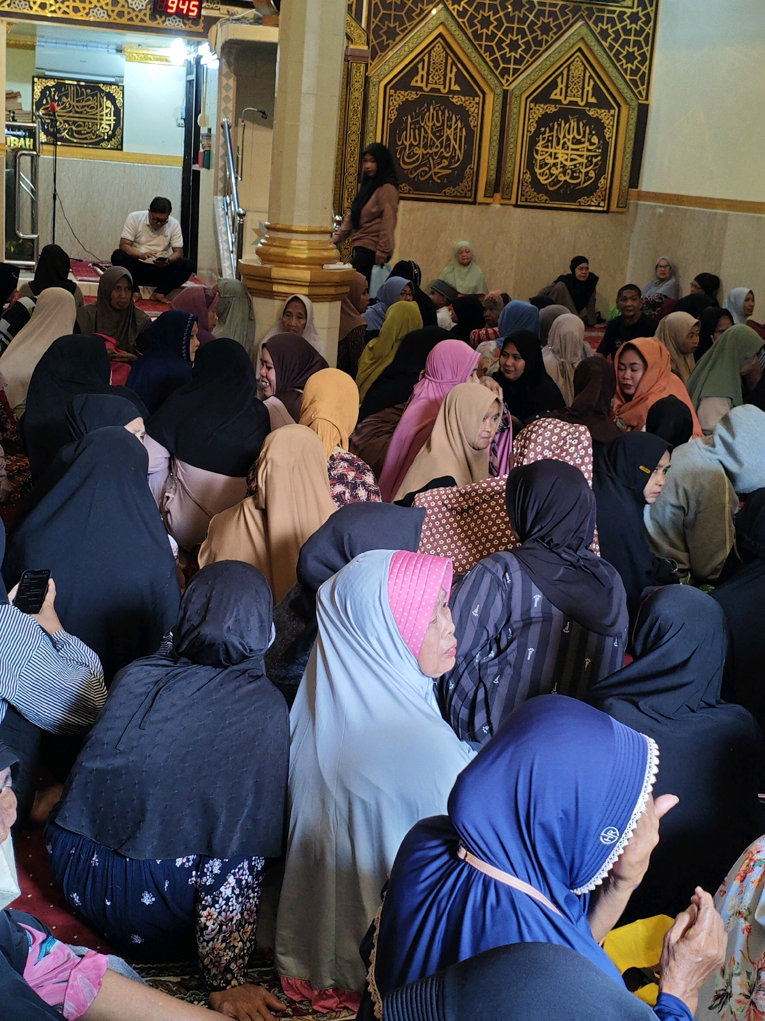 In Teddaopu, Indonesia, a group of people, predominantly women, are gathered indoors, likely in a mosque, as evidenced by the Islamic calligraphy on the wall and the presence of a man reading from a book in a semi-raised area. The room has a tiled floor and cream-colored walls. Decorative Islamic calligraphy panels are mounted on the wall. A digital clock showing "9:45" is visible in the background. Many individuals are wearing hijabs in various colors, including black, grey, pink, and light blue. Some are seated on the floor, while others appear to be standing or partially obscured. The overall atmosphere suggests a religious gathering or event.