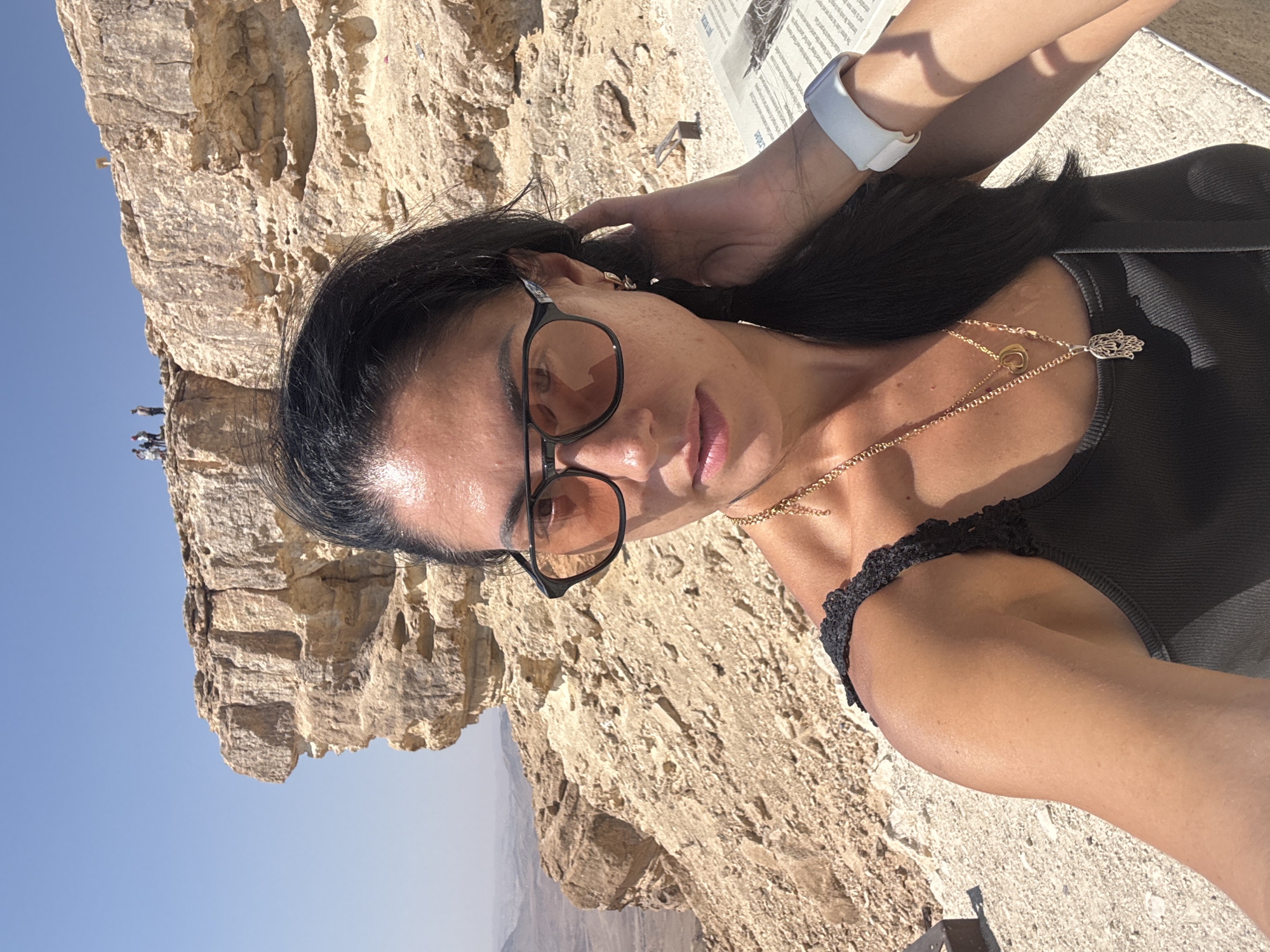 A woman with dark hair, wearing sunglasses and a black tank top with lace trim, takes a selfie at the Ramon Crater in Mitzpe Ramon, Israel. She is adorned with two gold necklaces, one featuring a crescent moon shape and the other a hamsa hand pendant. A white smartwatch is visible on her left wrist. The background consists of a vast, arid landscape with layered rock formations characteristic of the crater. In the distance, a group of people are visible standing on a cliff edge. The sky is clear and blue.