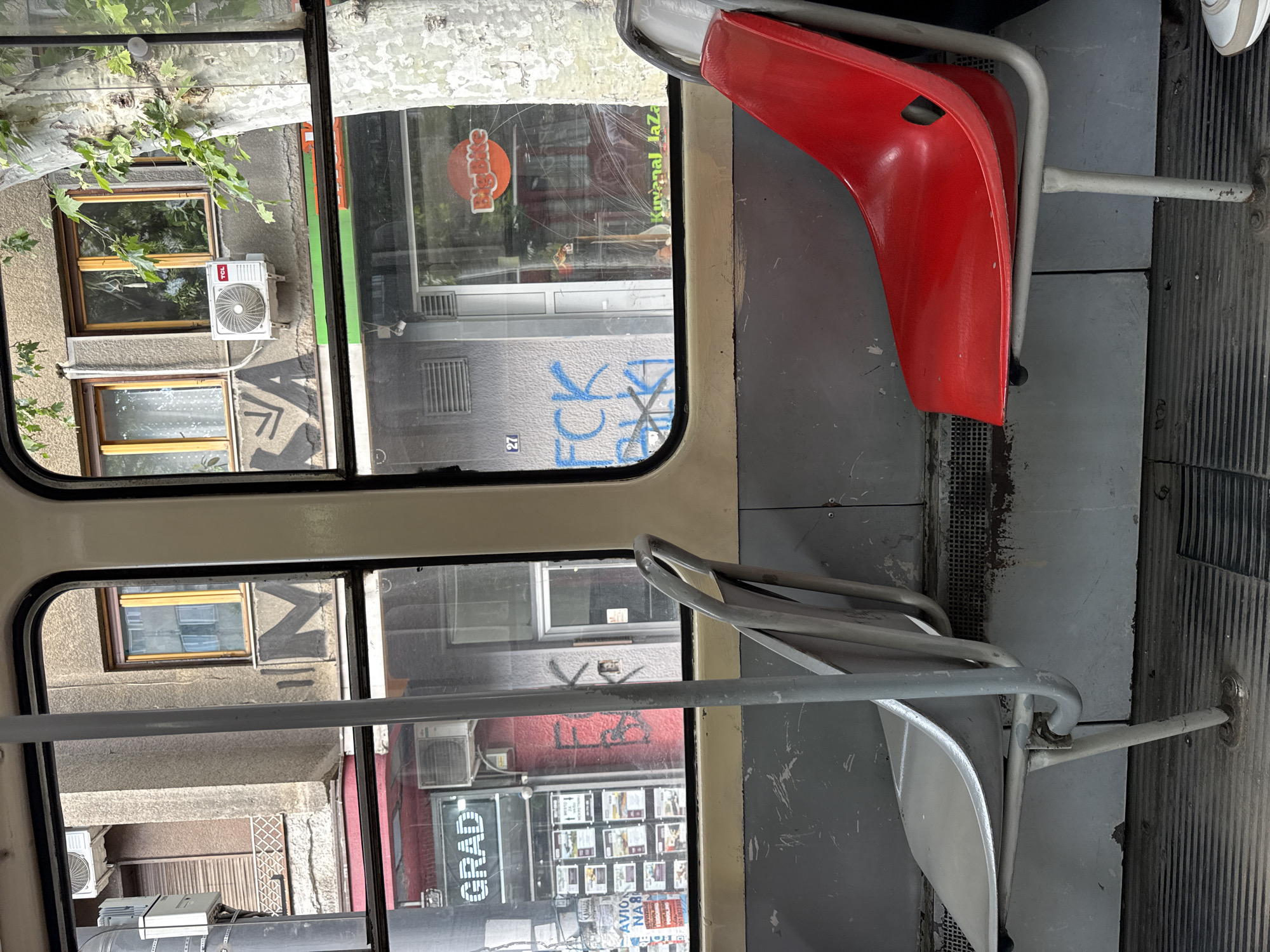 The photograph, taken from the interior of a public transport vehicle in Belgrade, Serbia, offers a view through the windows onto the city's architecture. The interior features grey and red plastic seats with silver metal frames. Outside the windows, one can see buildings with a variety of architectural styles, including older buildings with air conditioning units and graffiti. One window view includes a "Big Bite" restaurant sign and the address number 27 spray-painted on a building. Another window reveals a real estate agency advertising properties. Trees with green foliage are visible outside one window. The angle of the shot indicates the photograph was taken while the vehicle was in motion or stationary on a slight incline. A small portion of a shoe is visible.