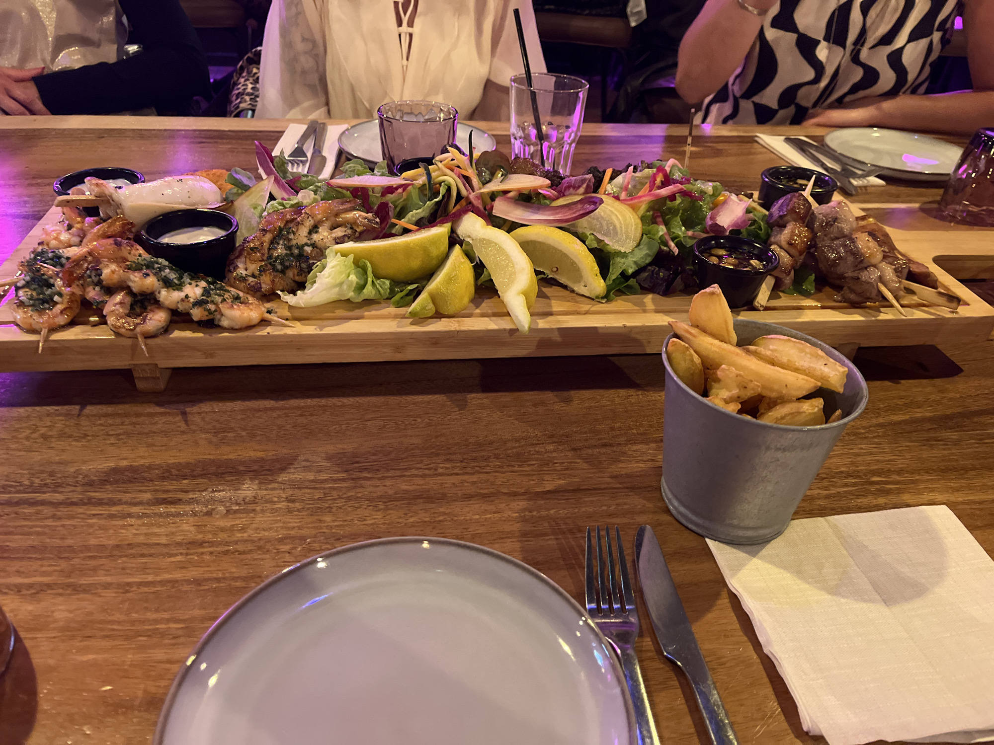 The image captures a dining scene in an unidentified location that appears to be a restaurant. A wooden table is set with various items. In the foreground, there's a plate, a fork, a knife, and a napkin. A metal bucket is filled with french fries. The table is dominated by a long wooden platter filled with assorted foods: grilled seafood skewers, chicken, lemon wedges, and a salad mix. Small black bowls contain sauces. In the background, three people are partially visible.
I do not have location information for Saint-Jean-de-Védas, France.
