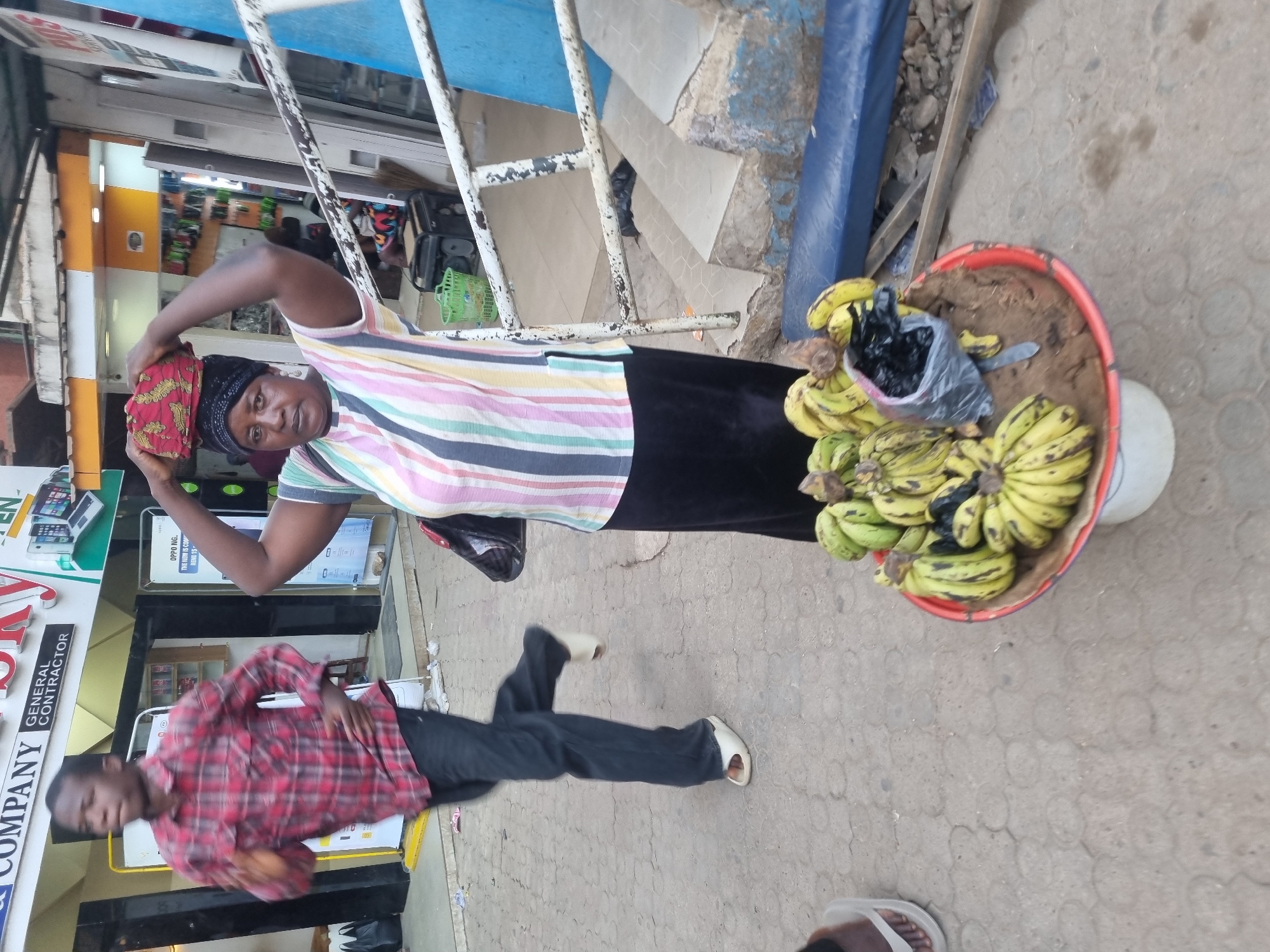 The image depicts a woman in a striped shirt and black skirt adjusting a head covering near a basket filled with bananas and a plastic bag containing something black. The location appears to be an outdoor street scene, potentially in Jos, Nigeria. The woman is standing near a metal railing. There is a building with a sign visible behind her, and a man in a red plaid shirt is lying on the ground in the foreground. The background shows parts of shops and street details.