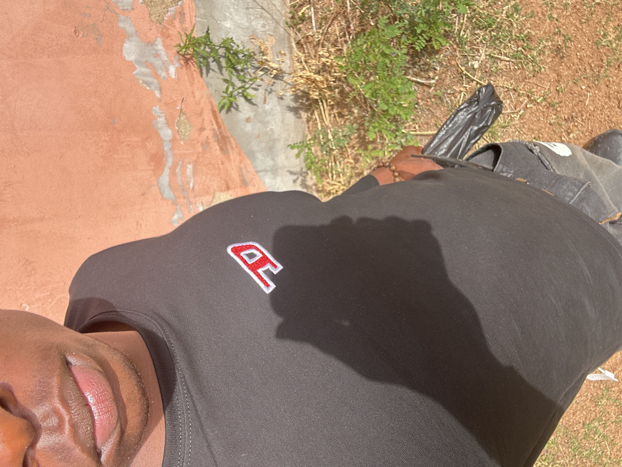 The image shows a close-up view of a person wearing a black t-shirt with a red "R" logo. Part of the person's face, specifically their lips and chin, are visible in the lower-left corner. The background includes a concrete surface with peeling orange paint, some sparse green vegetation, and a patch of dry, brown earth. A black plastic bag and what appears to be a dark-colored bag or backpack are also visible in the background. The image doesn't provide location information or specific details related to the city of Kirana, Nigeria.