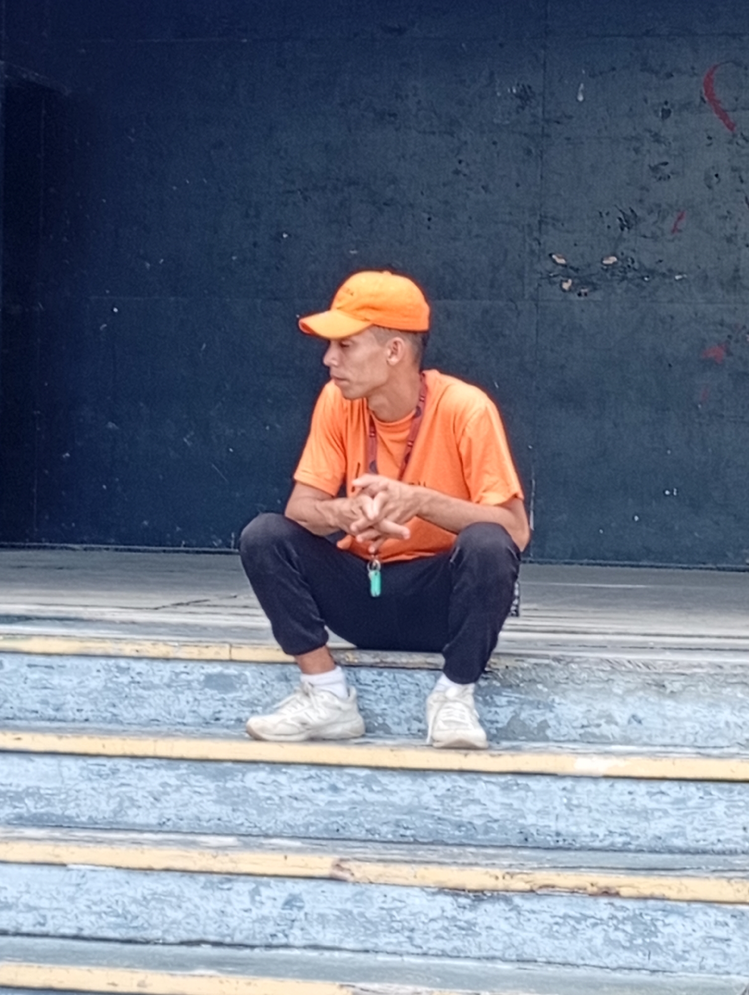 A man wearing an orange t-shirt and cap is squatting on a set of weathered, blue and yellow steps in Playa El Agua, Venezuela. He is wearing dark pants and white shoes, and has a lanyard with a green object attached around his neck. The background is a dark, textured wall.