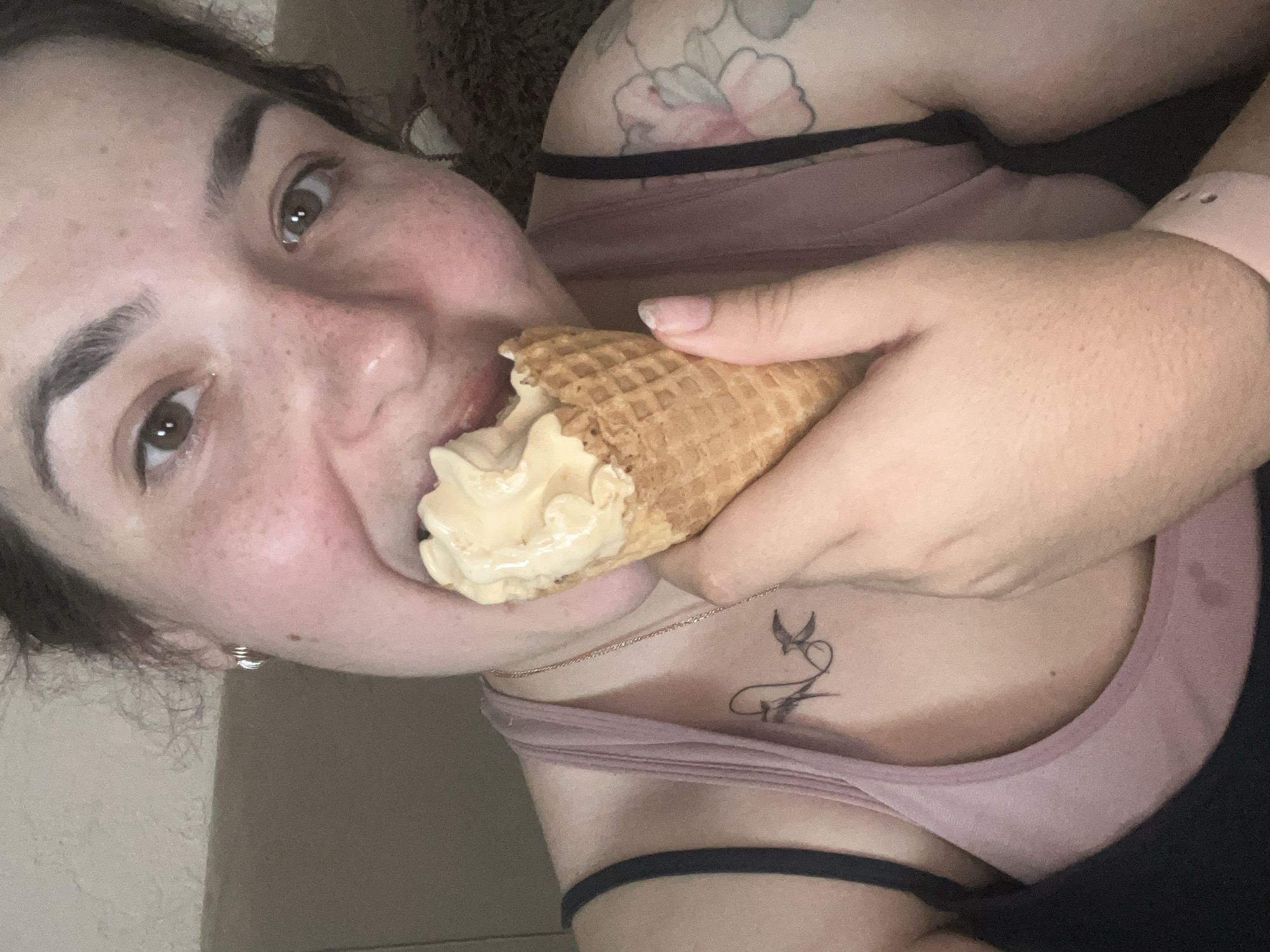 The image shows a close-up of a woman eating an ice cream cone. She is wearing a pink tank top and has freckles on her face. There are tattoos visible on her chest and arm. The background is out of focus and appears to be indoors. The scene is set in Krasnyye Barrikady, Russia.