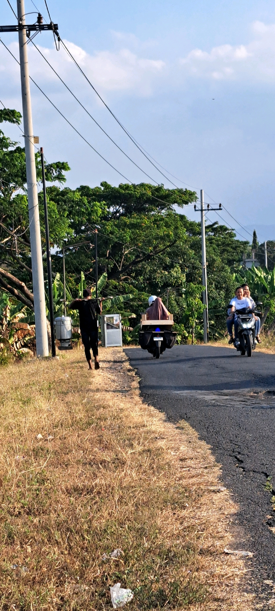 The image depicts a scene on a road in Kebon Waris, Indonesia, with a person walking along the side and two motorcycles on the road. A man in a black shirt and pants is walking on the grassy shoulder of the road, carrying a pole over his shoulders with two large containers attached, one on each side. He is barefoot and walking away from the viewer. To his right, on the asphalt road, a motorcycle is carrying two people. The person in the front is wearing a white shirt and jeans and appears to be smiling. The person behind is wearing a white helmet and a brown garment covering their torso and head. Further down the road, another motorcycle is visible, carrying one person with a white helmet and a brown garment. This motorcycle has a large wooden box strapped to the back. The environment includes a utility pole with wires, lush green trees in the background, and dry, yellow grass along the side of the road. The sky is blue with some scattered clouds.