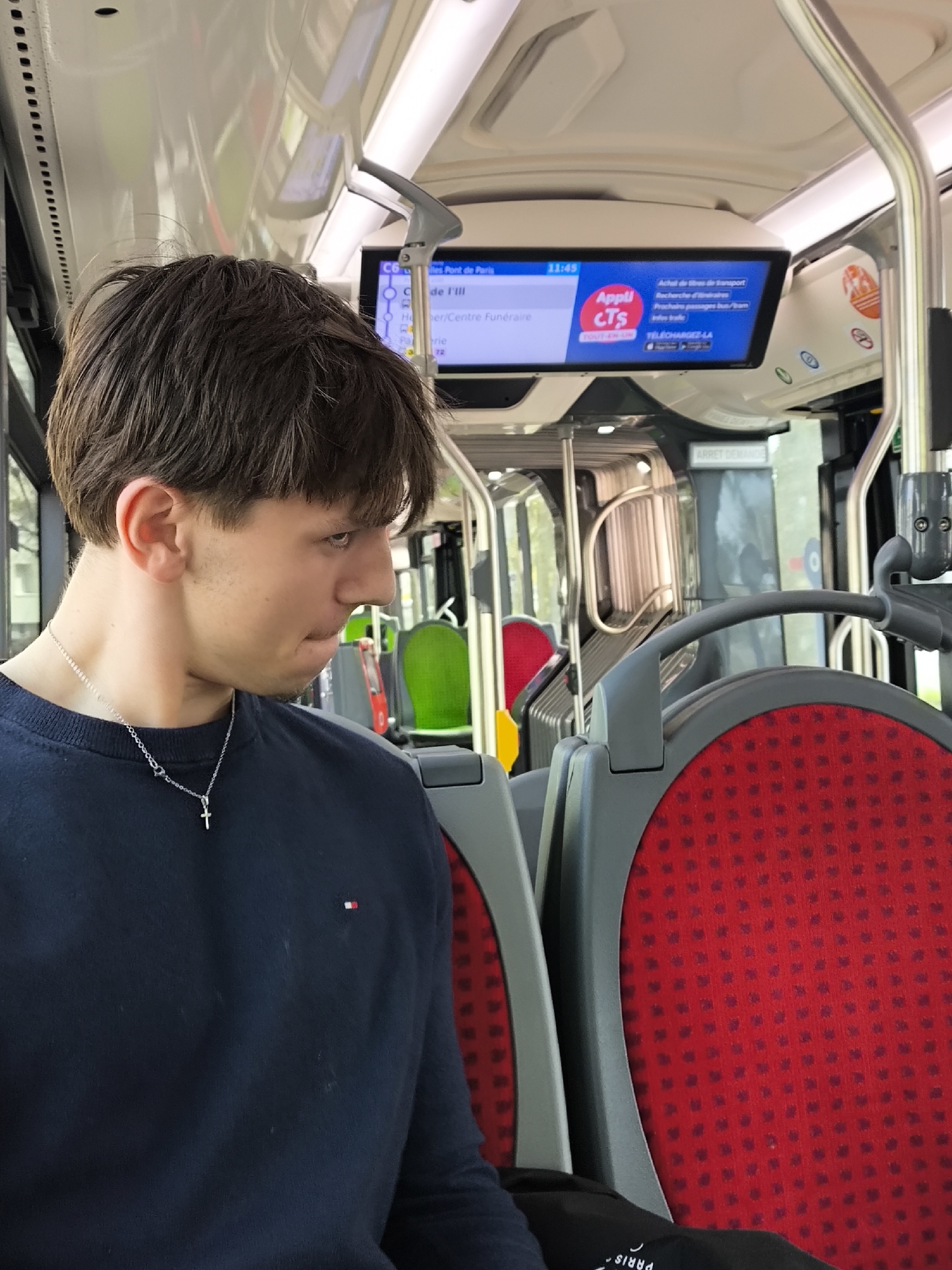 Here's a detailed description of the image:
**Scene:** Interior of a CTS (Compagnie des Transports Strasbourgeois) bus in Strasbourg, France. The image is taken from the perspective of a passenger, likely seated behind and to the left of the main subject.
**Subject:** A young man with dark, short hair and a silver cross necklace. He is wearing a dark blue crew neck shirt with a small red and white logo. He appears to be looking to his left with a slight frown or pensive expression.
**Objects:**
* Bus seats: The foreground shows a red patterned bus seat with a gray frame. Other seats with red, green, and yellow backs are visible further inside the bus.
* CTS information screen: A digital display is mounted on the bus ceiling, showing the current bus route ("C6 les Pont de Paris"), next stops ("Île I'III"), a transportation app logo, and information about purchasing tickets.
* Handrails and poles: Gray metal poles and handrails are visible throughout the bus interior.
* Bus windows: The windows show an out-of-focus glimpse of the outside environment.
**Environment:**
The bus interior is well-lit. The overall setting is a public transportation vehicle traveling within the city of Strasbourg, France.
**Actions/Interactions:**
The image captures a static moment, with the young man seemingly lost in thought or observing something outside the frame. The bus appears to be in transit based on the information displayed on the screen.